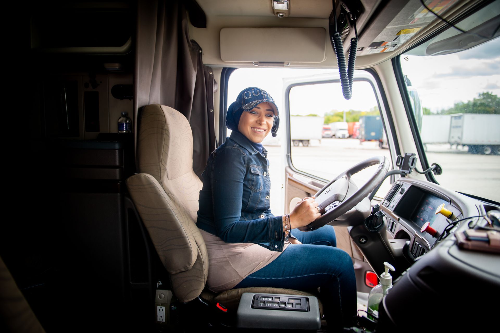 A smiling person in a denim jacket and cap sitting in the driver’s seat of a parked semi-truck.