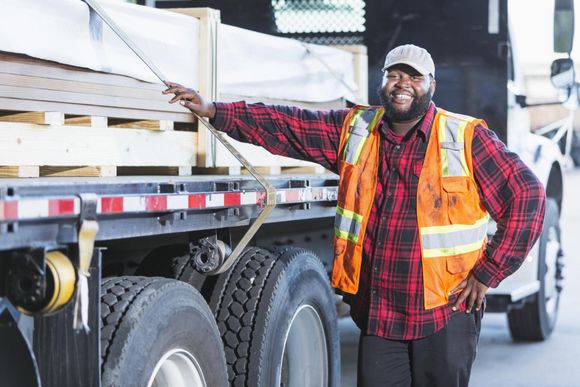 A man in a plaid shirt and orange vest is standing next to a semi truck.