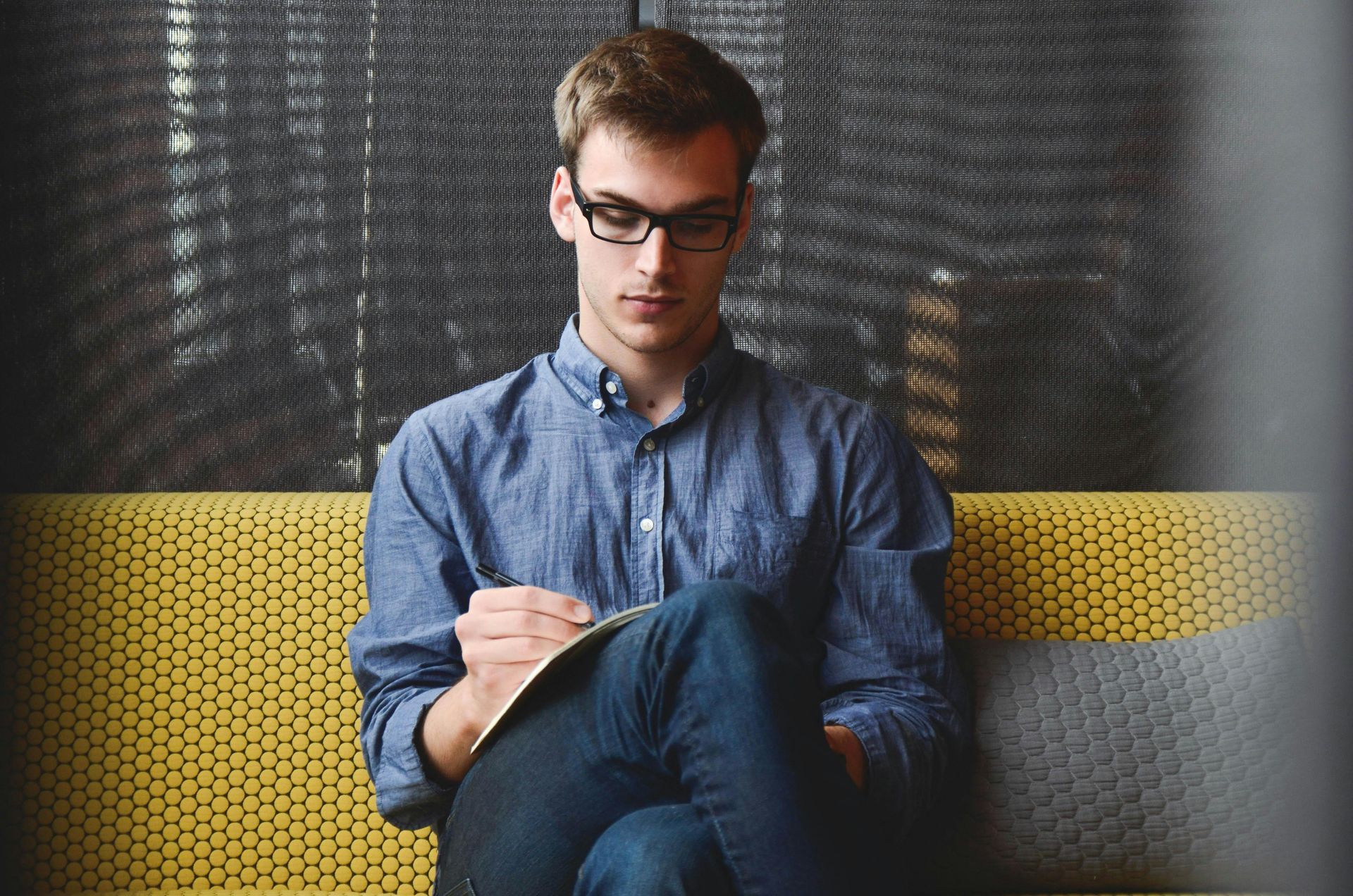 A person with glasses and a blue shirt sits on a yellow couch, focused on writing in a notebook.