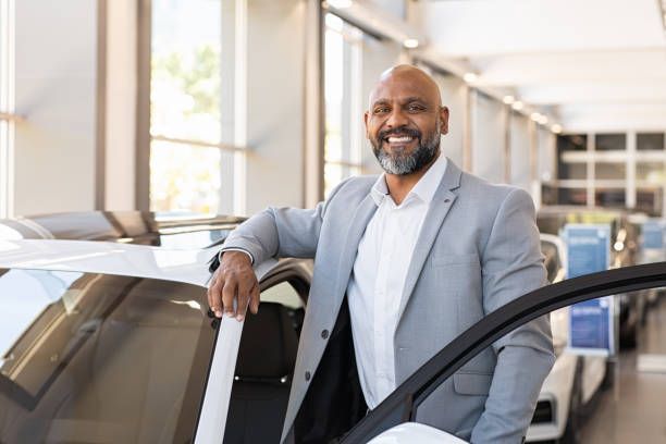 A man in a suit is standing next to a car in a showroom.