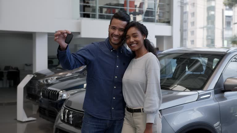 A man and a woman are standing in front of a car dealership holding a car key.