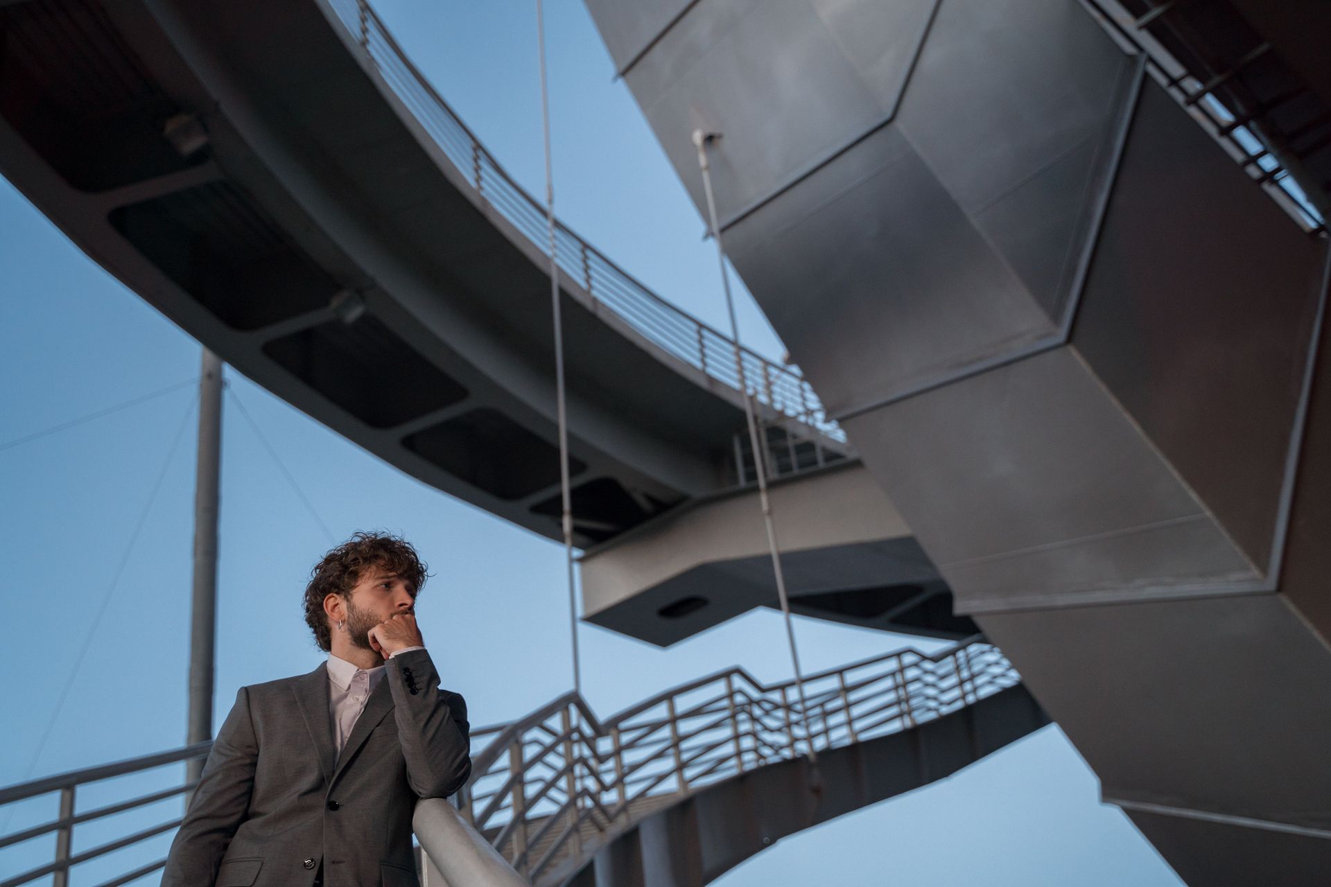 Person stands under a modern overpass, looking up beside metal stairs against a blue sky.