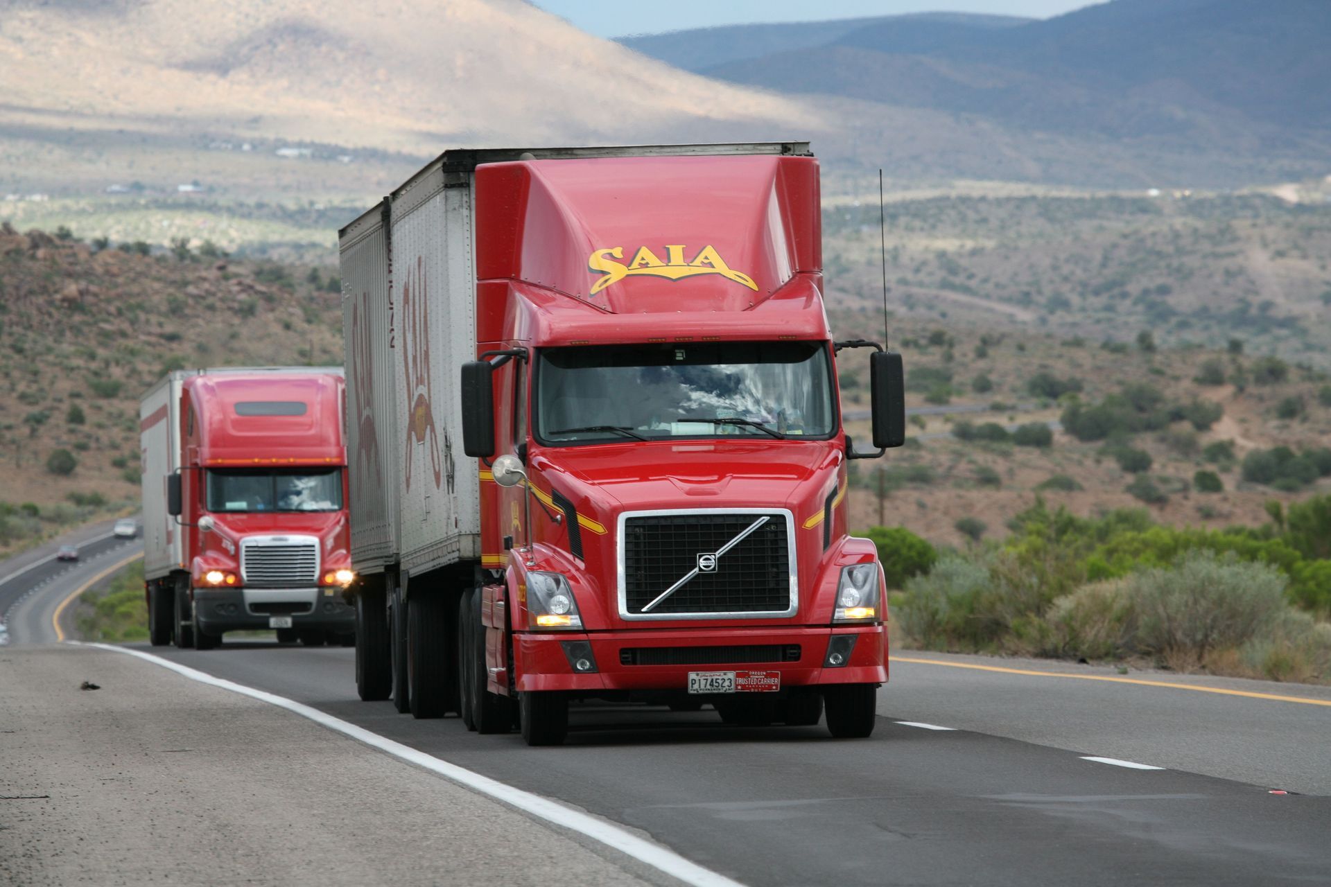 Two red Volvo semi trucks driving along a curving highway in a desert landscape