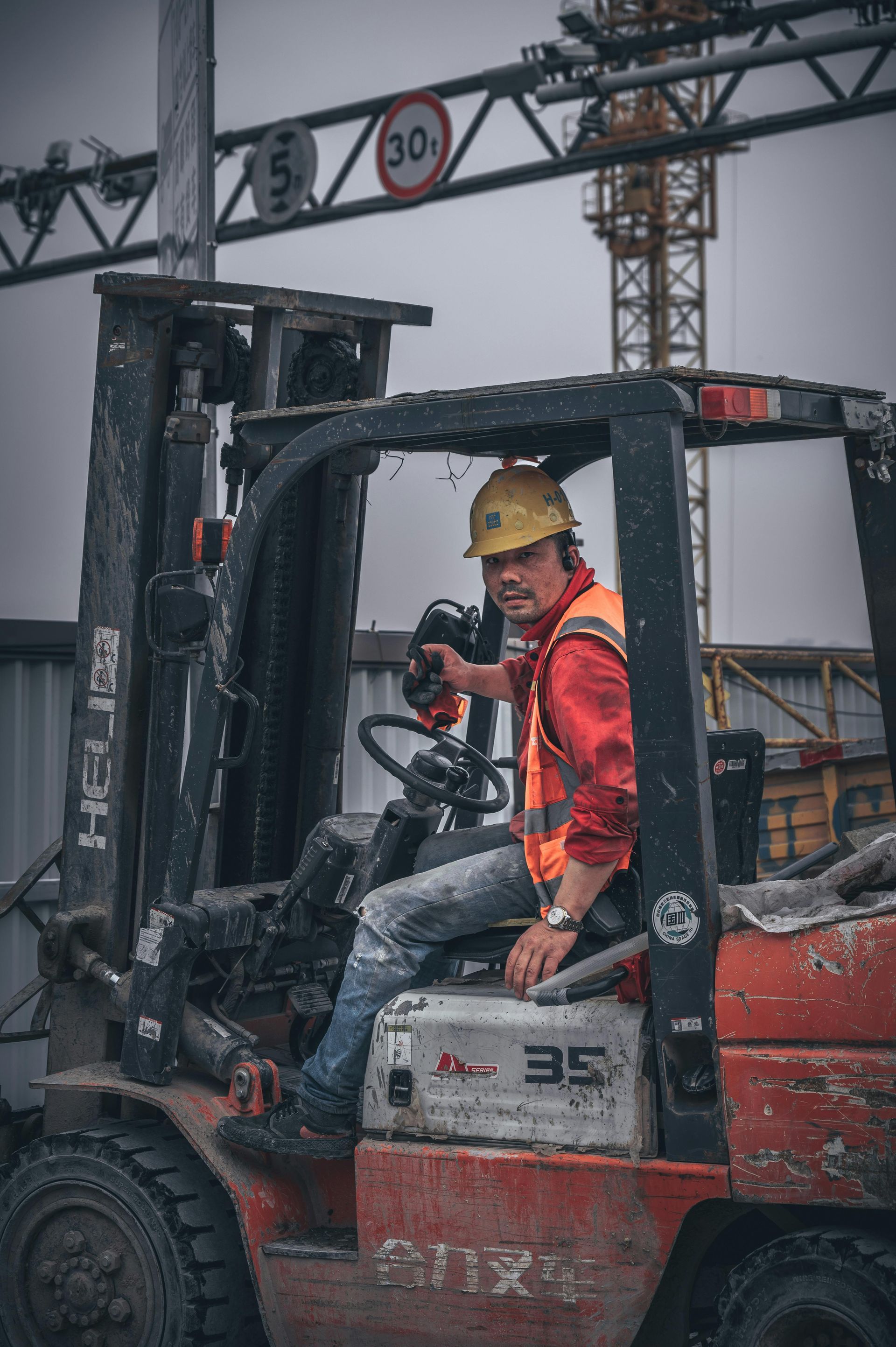 Construction worker in orange vest and hard hat sits in forklift on a job site.