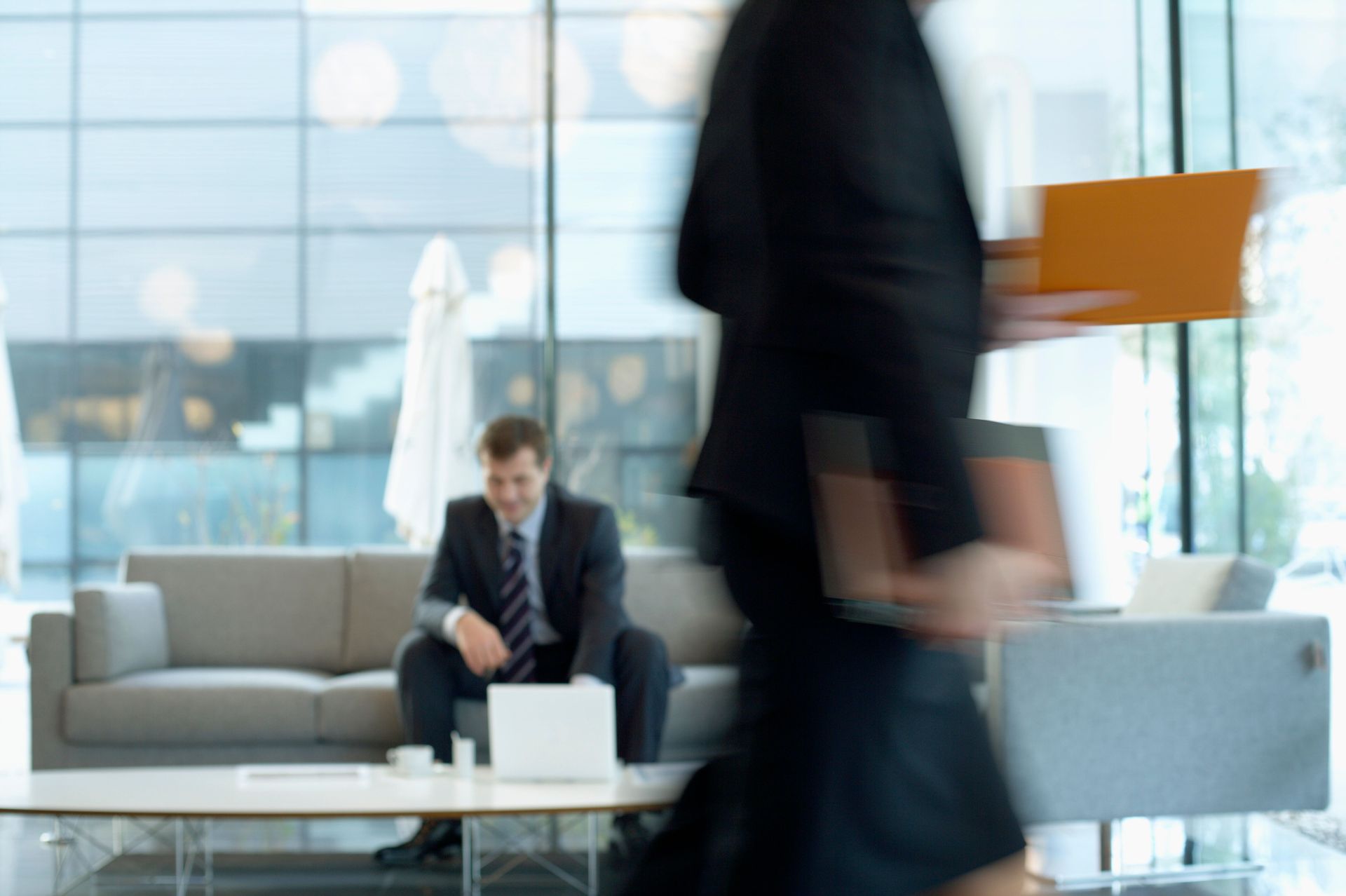 Man sitting on a couch in a modern office lobby while a person walks past in the foreground