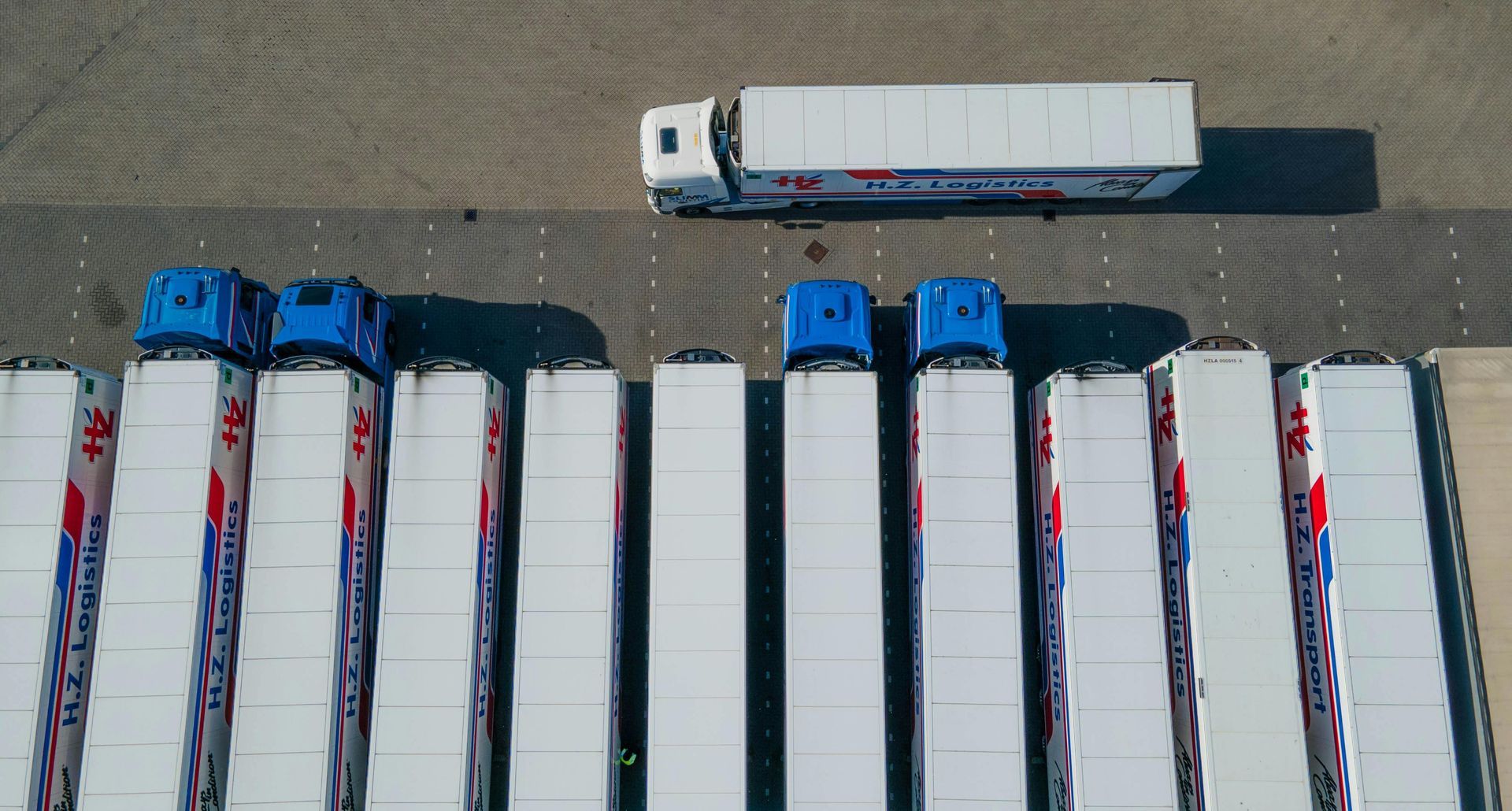 An aerial view of a row of blue-cabbed semi-trucks parked parallel to one another, with a white truck parked above them.