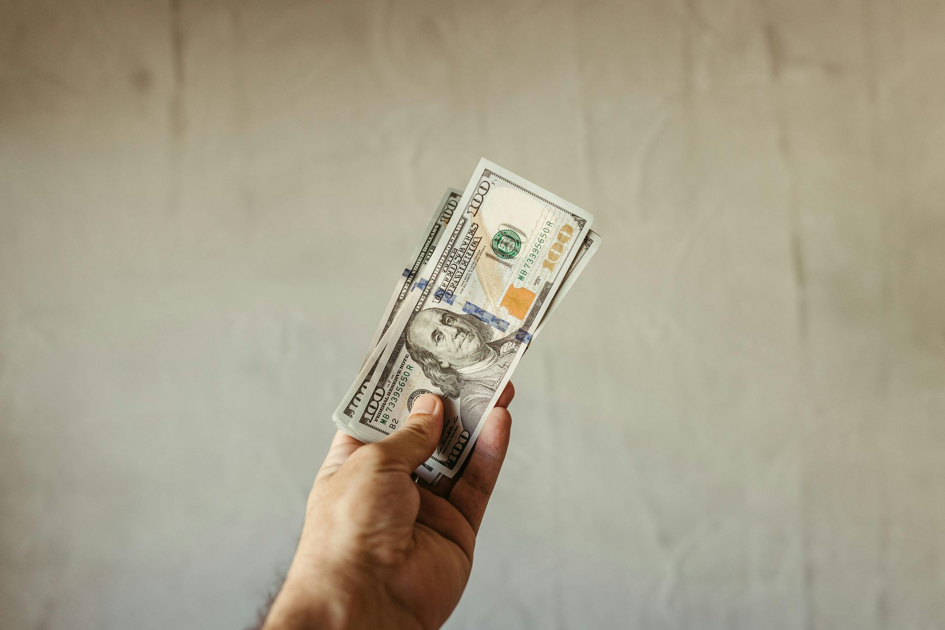 A hand holds several American hundred-dollar bills against a plain, light-colored background.