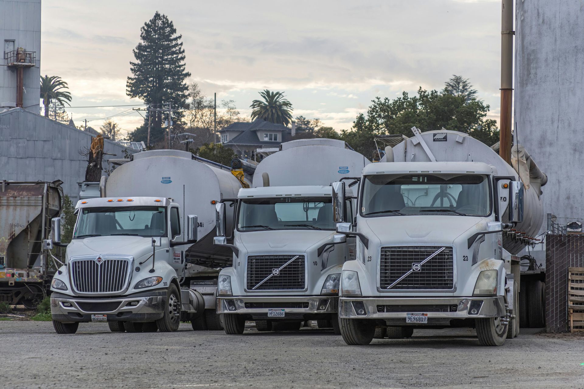 Three tanker trucks parked near industrial buildings, with overcast sky.