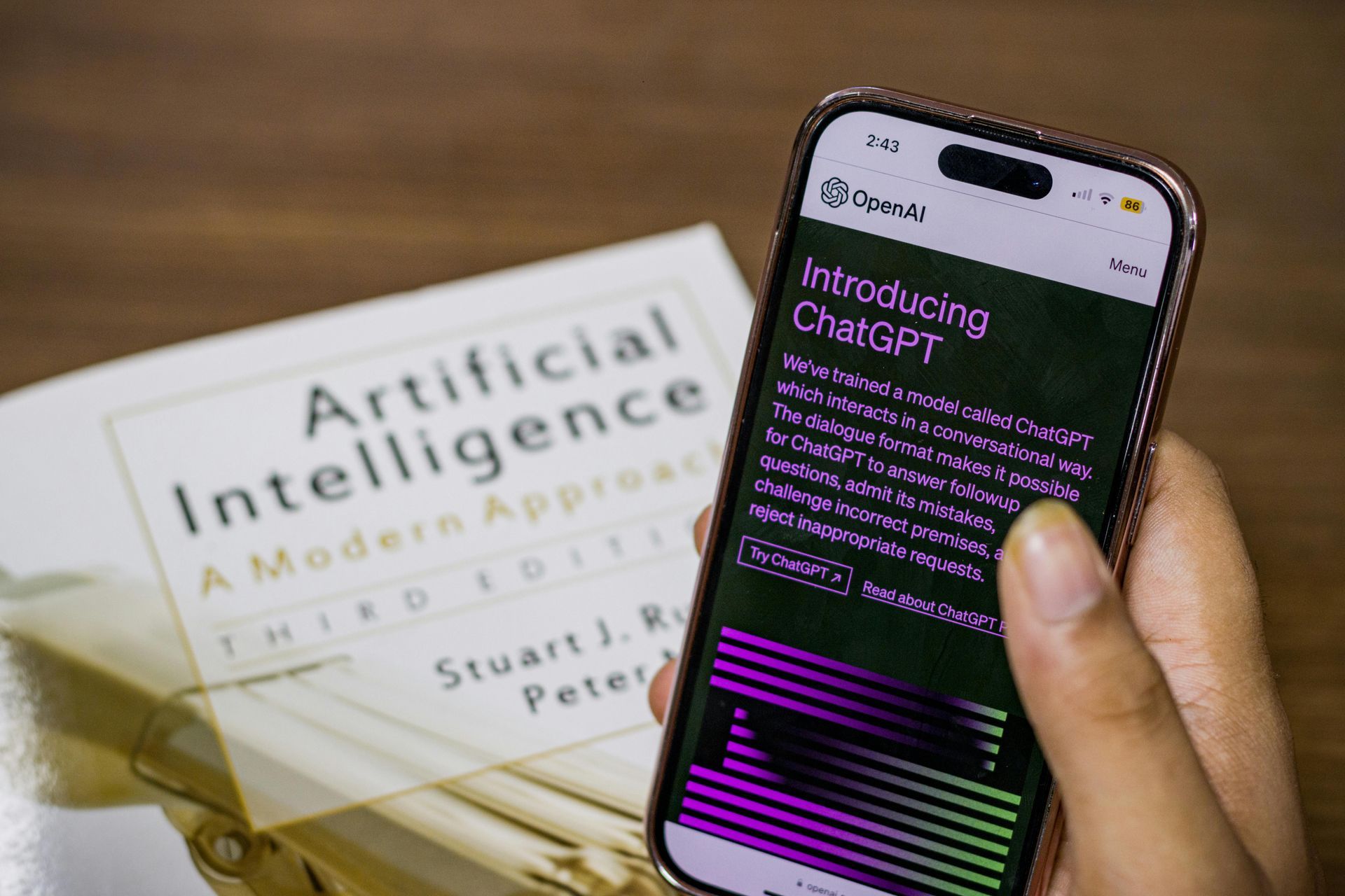A hand holds a smartphone displaying the ChatGPT introduction page next to an Artificial Intelligence textbook.