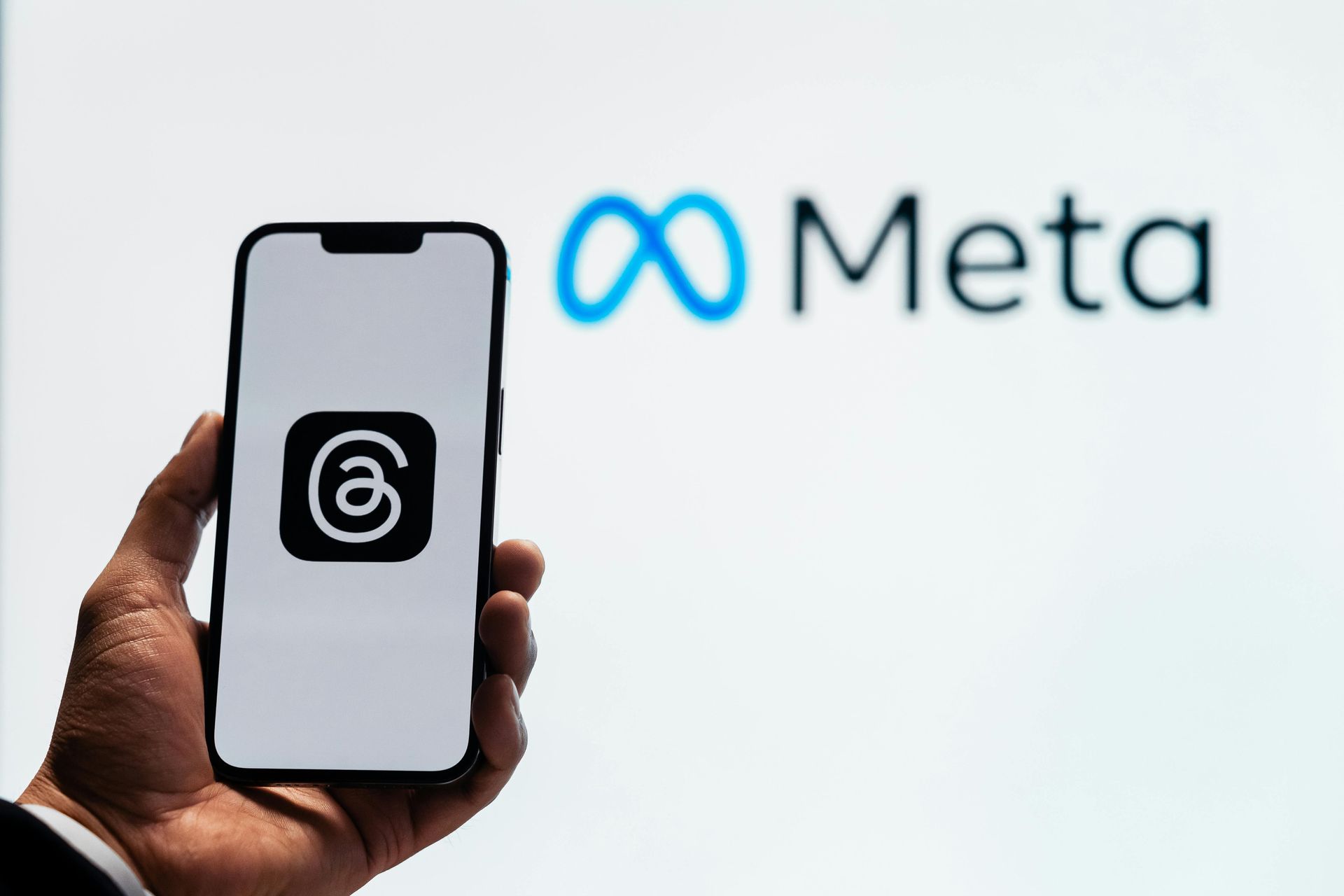 A hand holds a smartphone displaying the Threads logo in front of a blurred Meta logo.