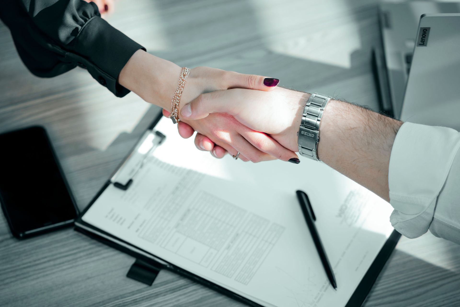 Two people shake hands over a desk with paperwork and a pen, suggesting a business agreement.