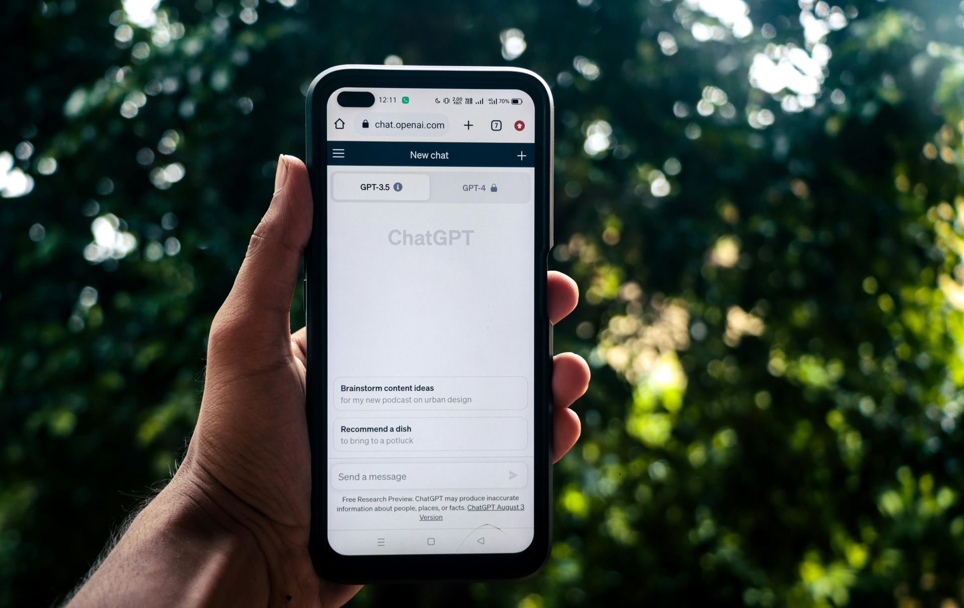 A hand holds a smartphone displaying the ChatGPT website against a blurred, leafy green background.
