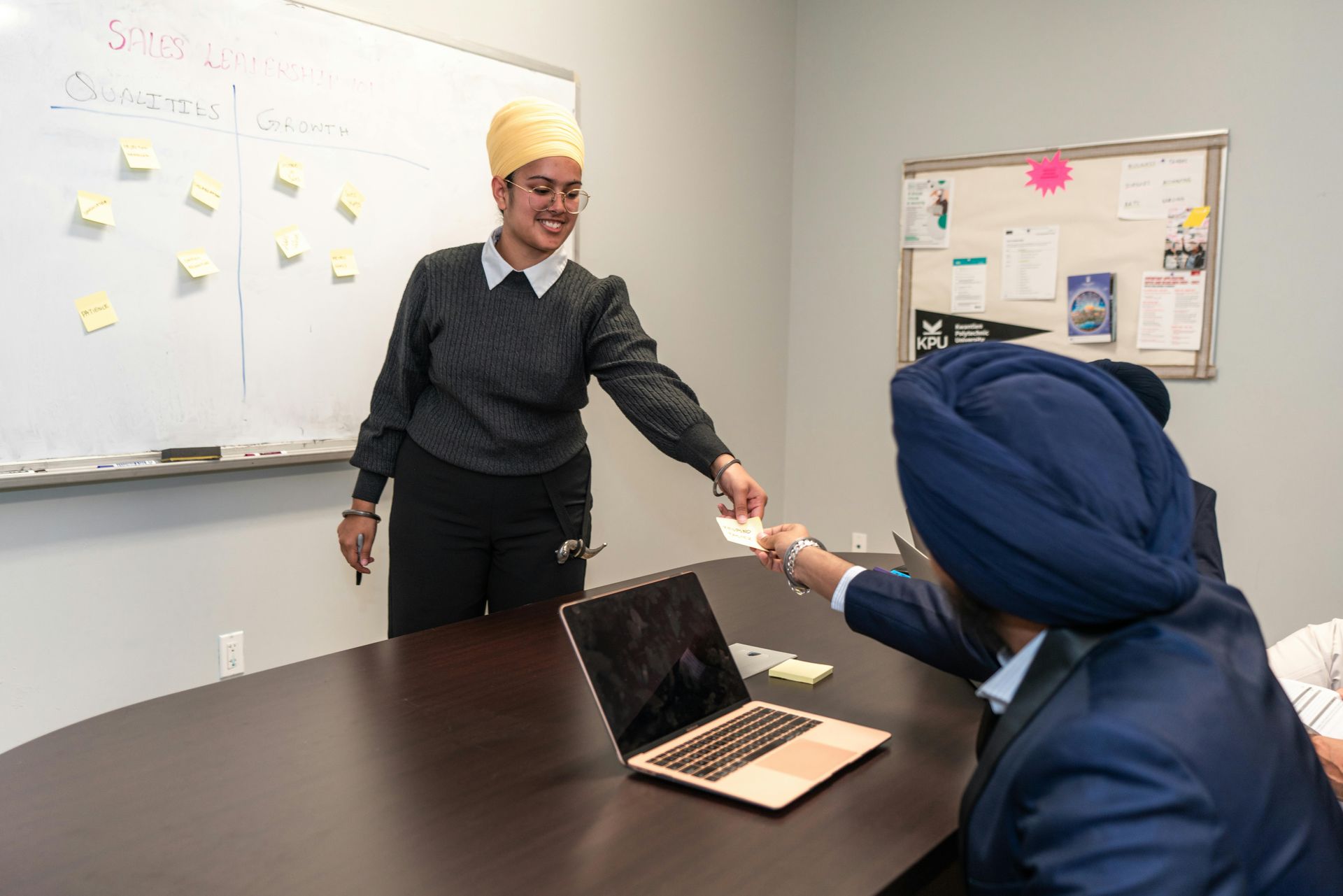 A person standing by a whiteboard with sticky notes hands a note to a colleague sitting at a table with a laptop.