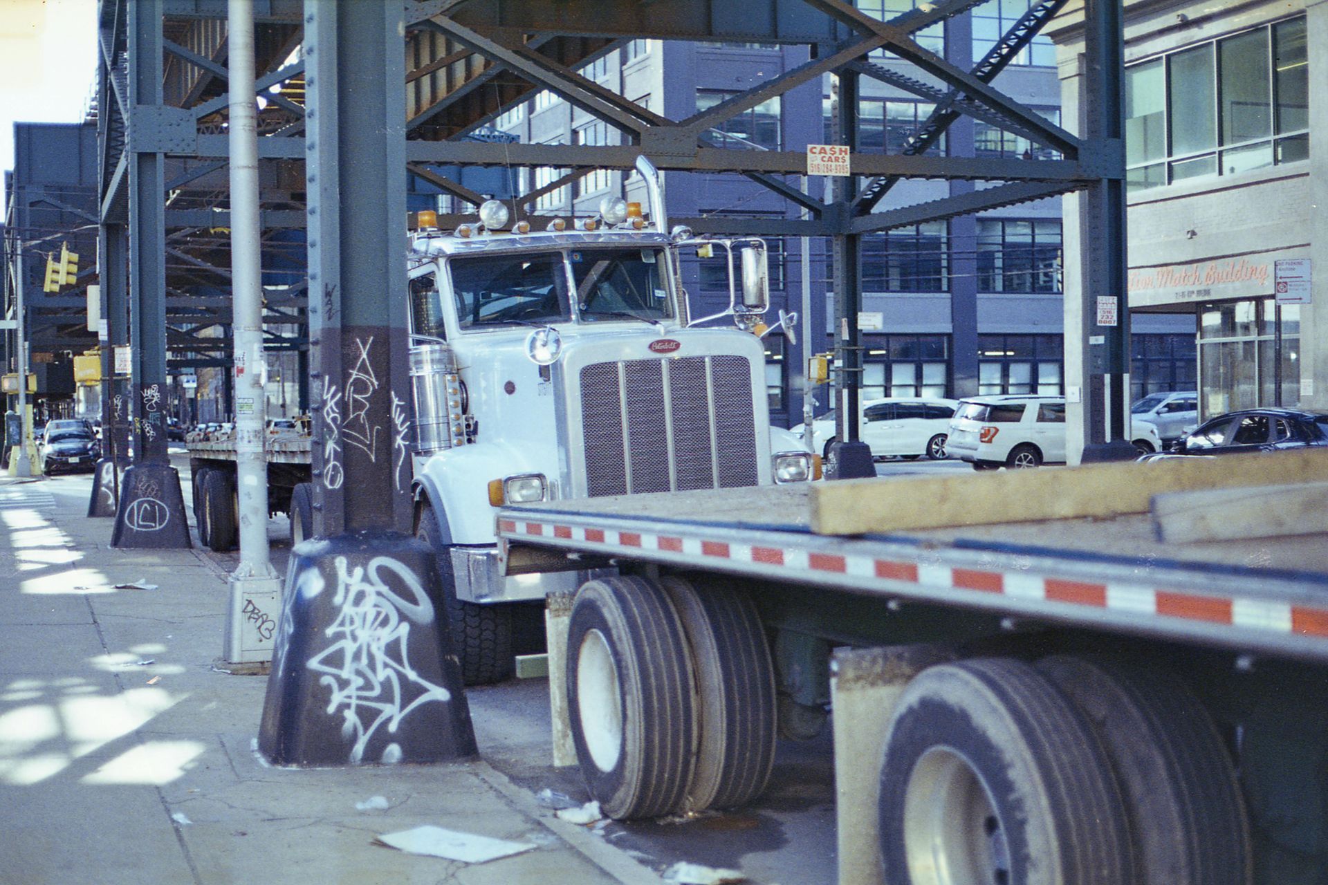 White semi-truck parked under an elevated train structure in a city, with graffiti on support beams.