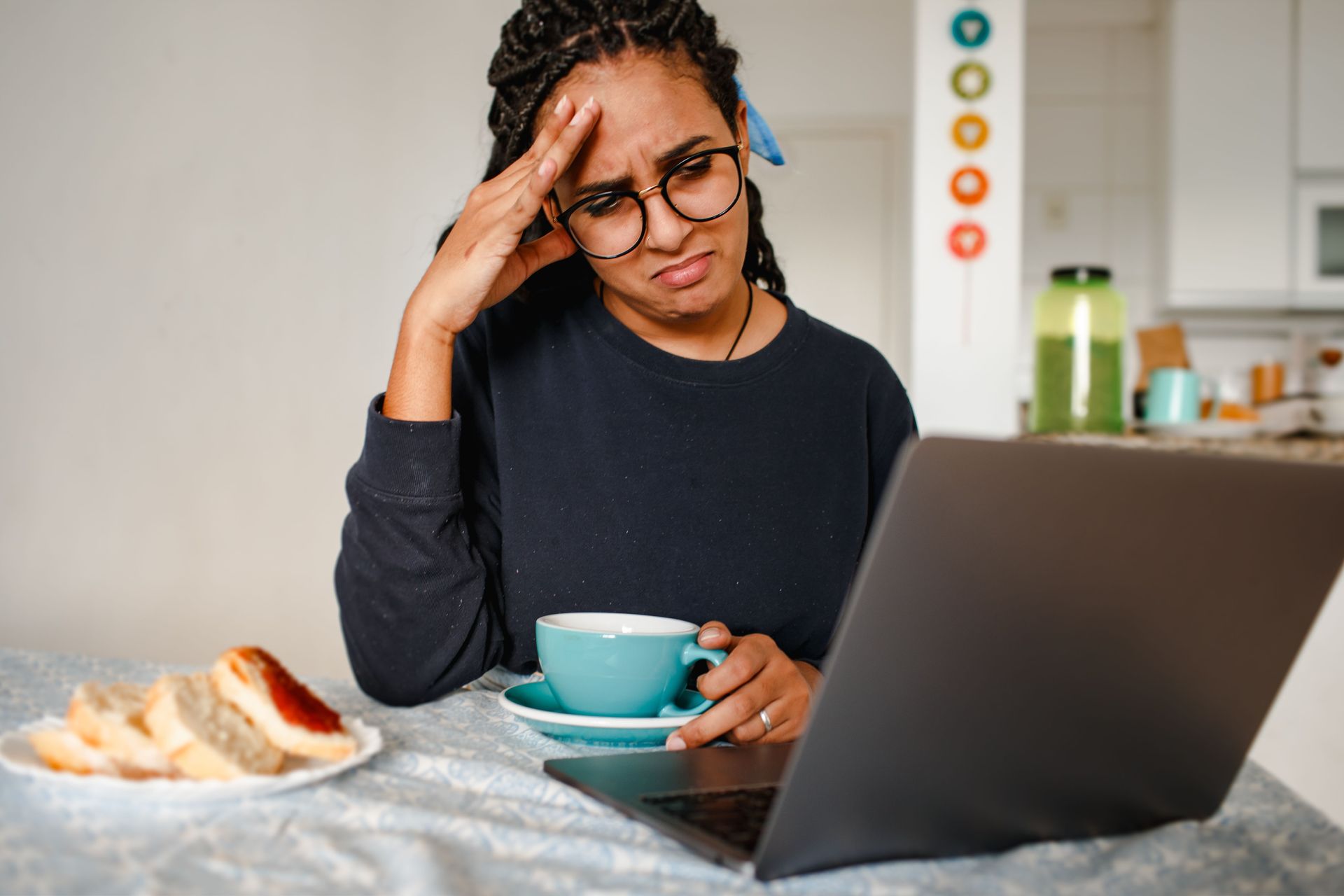 Person at a kitchen counter holding a mug and looking worried at a laptop, with bread nearby.