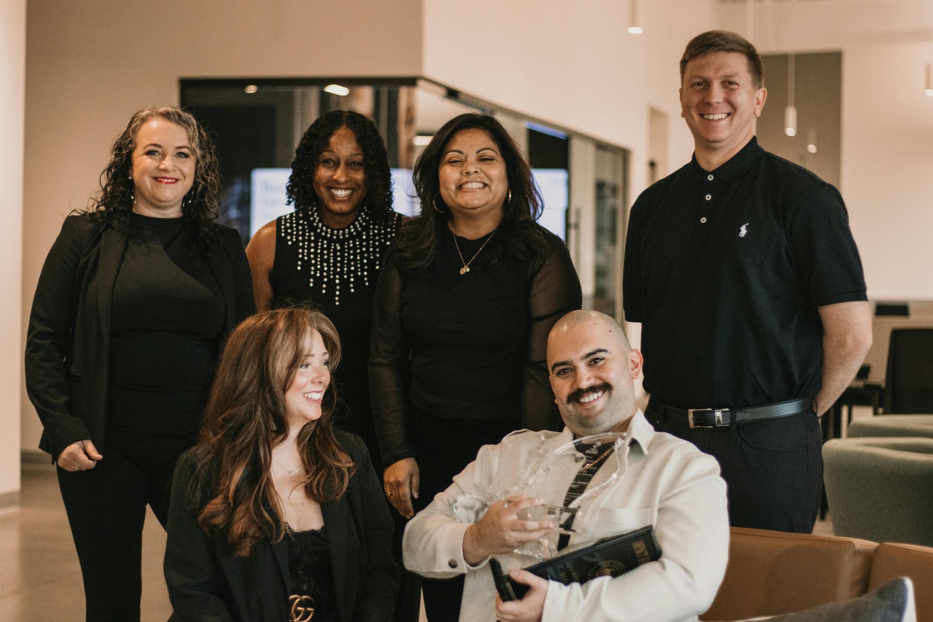 Group of six people smiling in an office setting, wearing business attire.