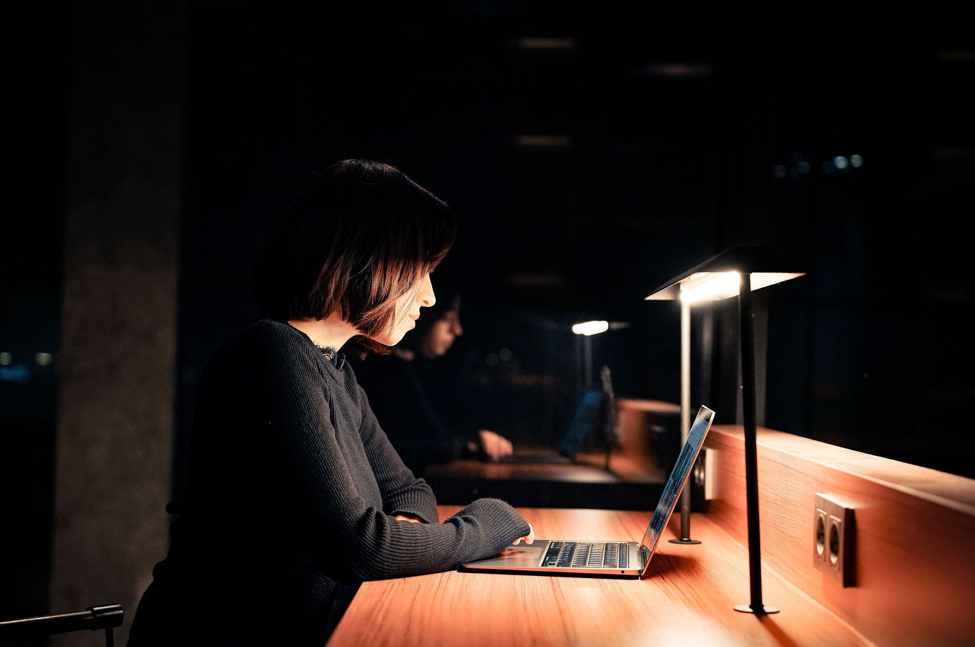Person typing on a laptop at a dimly lit desk under a bright lamp.