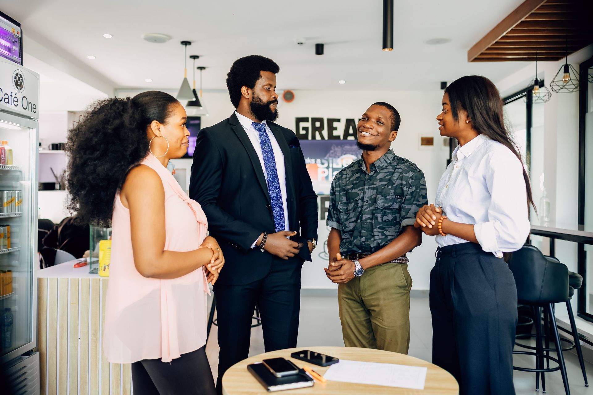 Four coworkers chatting in a bright office lobby, with a table and wall sign in the background.
