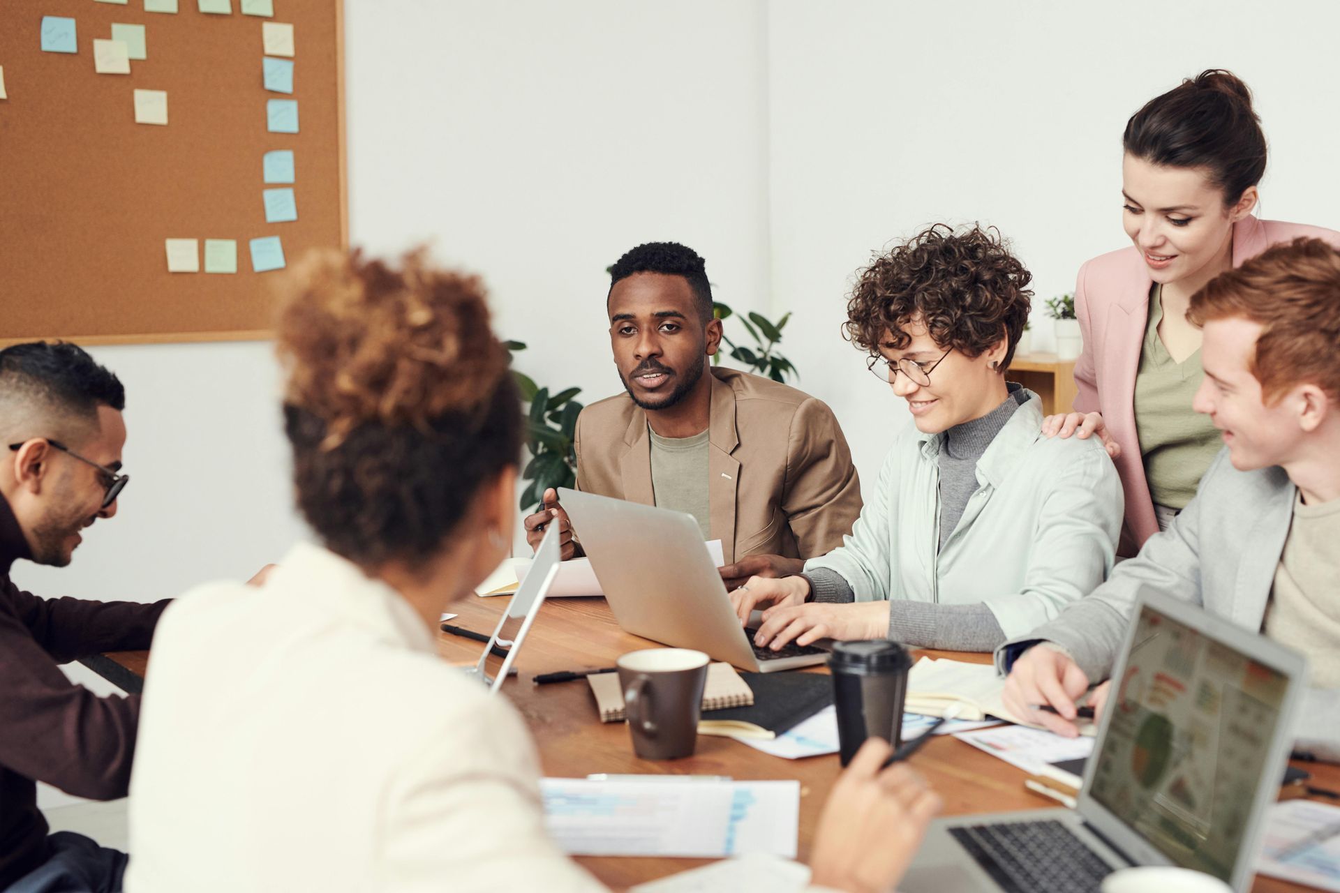 A diverse group of professionals collaborates around a wooden table in an office, using laptops and discussing work.