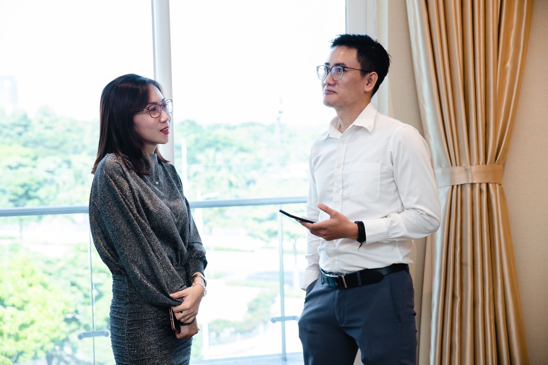 Two people talking indoors by a bright window, one holding a tablet and wearing a white shirt.
