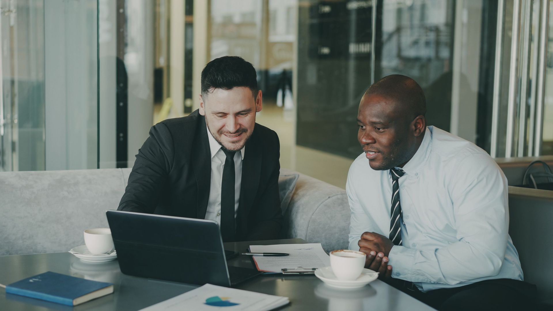 Two coworkers review a document together at a table with a laptop and coffee in a glass-walled office