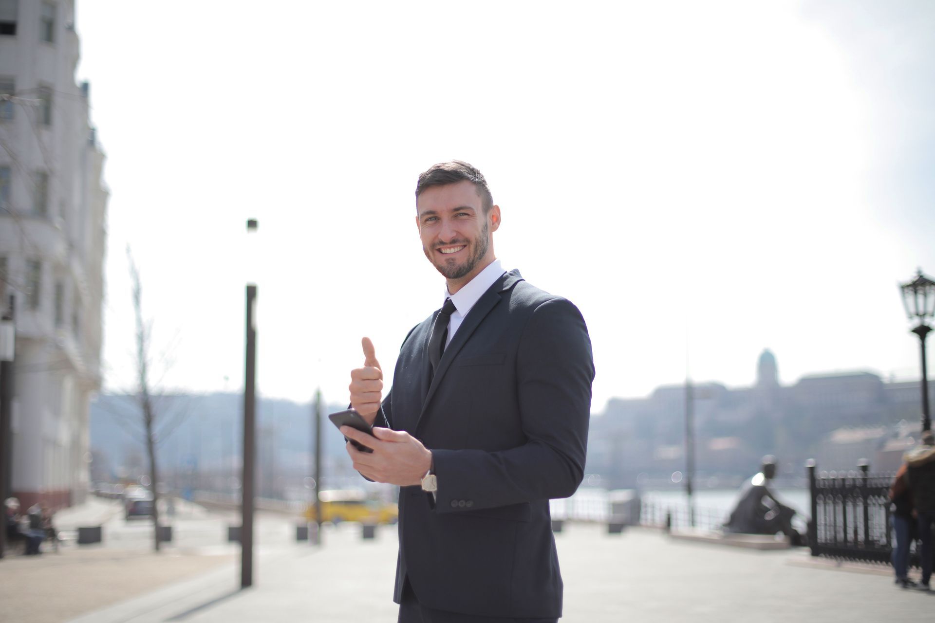 Smiling man in a dark suit holding a tablet on a waterfront promenade in bright daylight