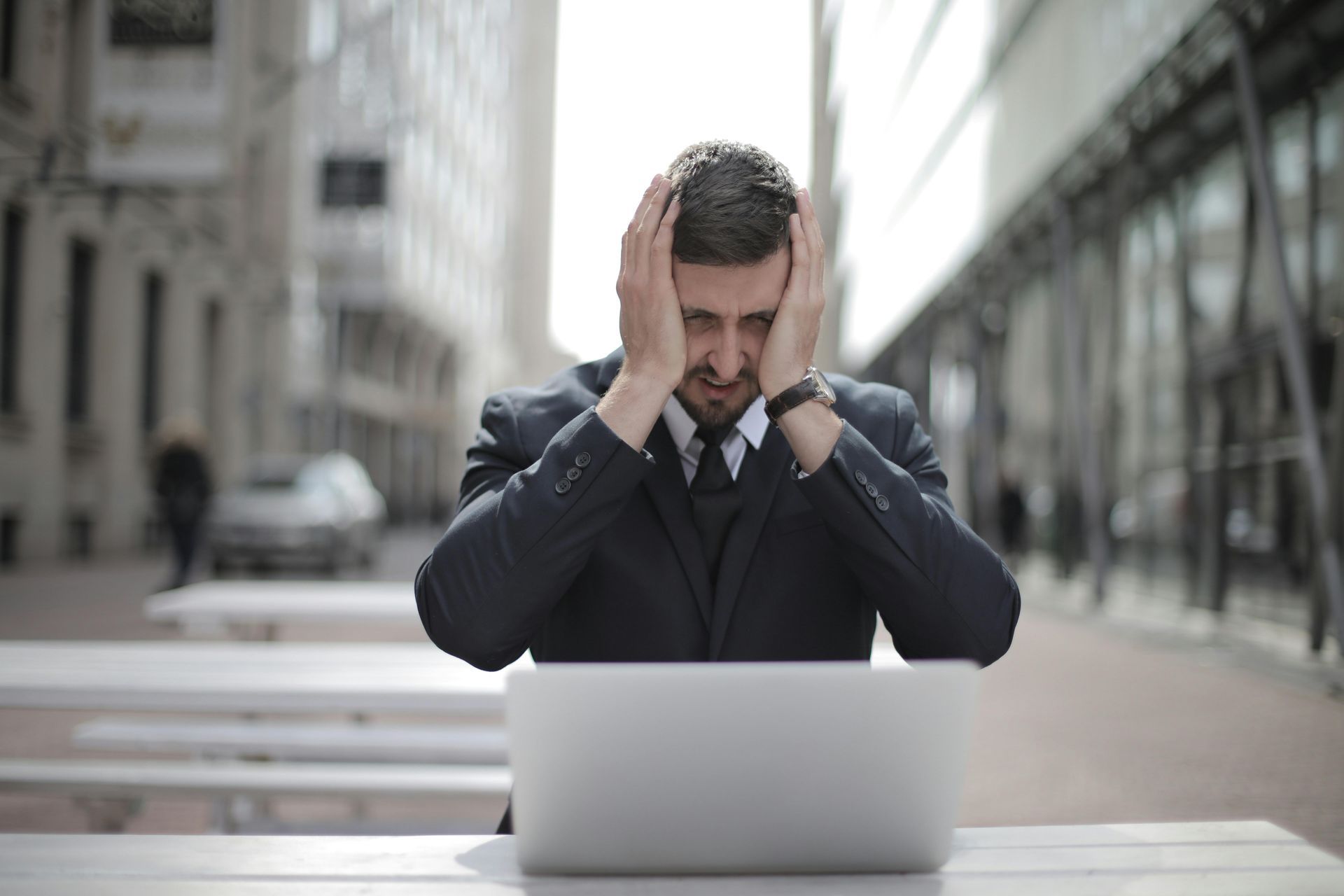 A businessperson in a suit sits at a laptop outdoors, holding their head with both hands in a look of frustration.