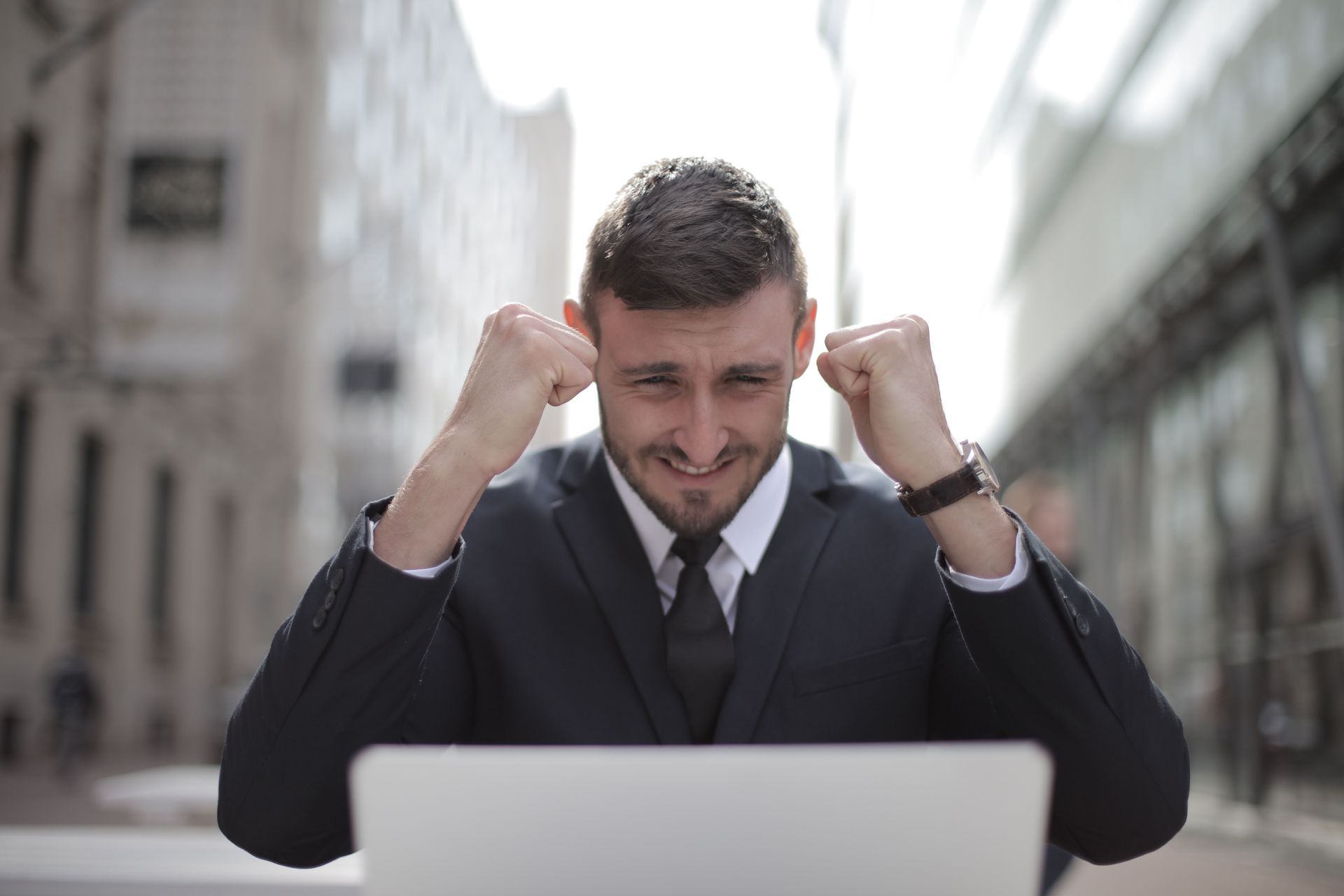Frustrated man in a suit clenching fists at a laptop outdoors in a city setting