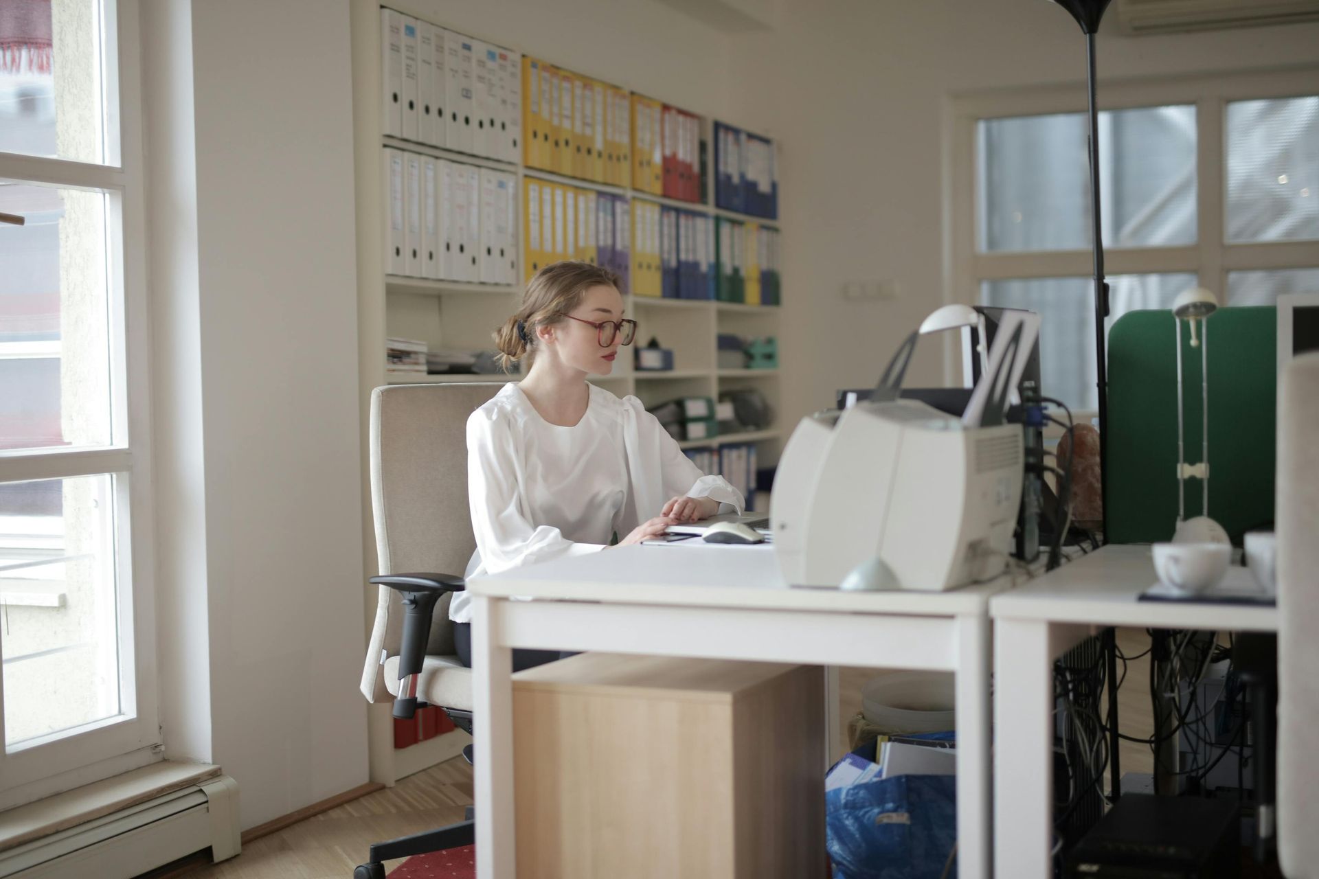 Woman working at a desk in a bright office, surrounded by shelves of files and a window.