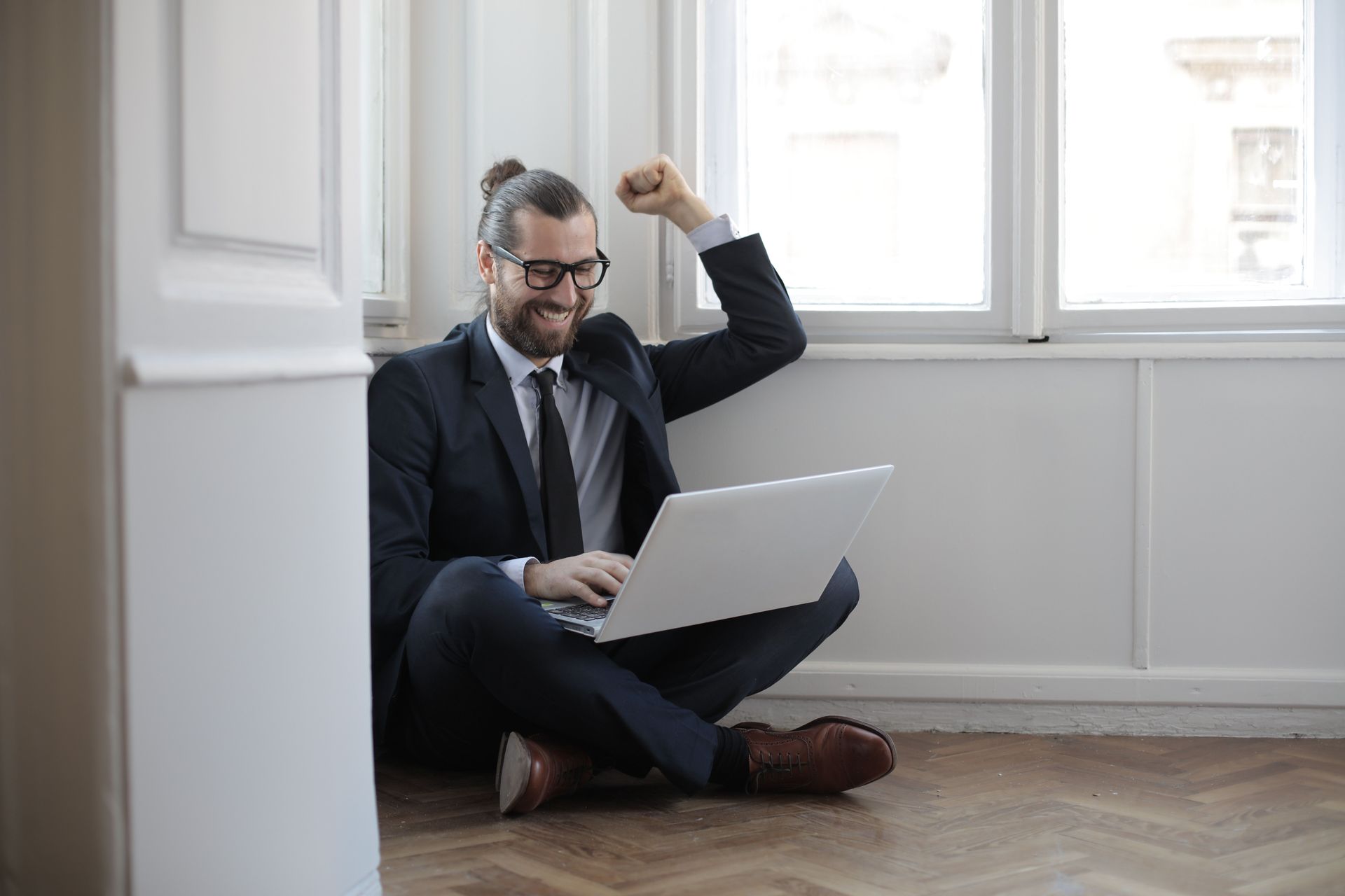 Man sitting on the floor with a laptop, raising a fist in a bright room by a window.