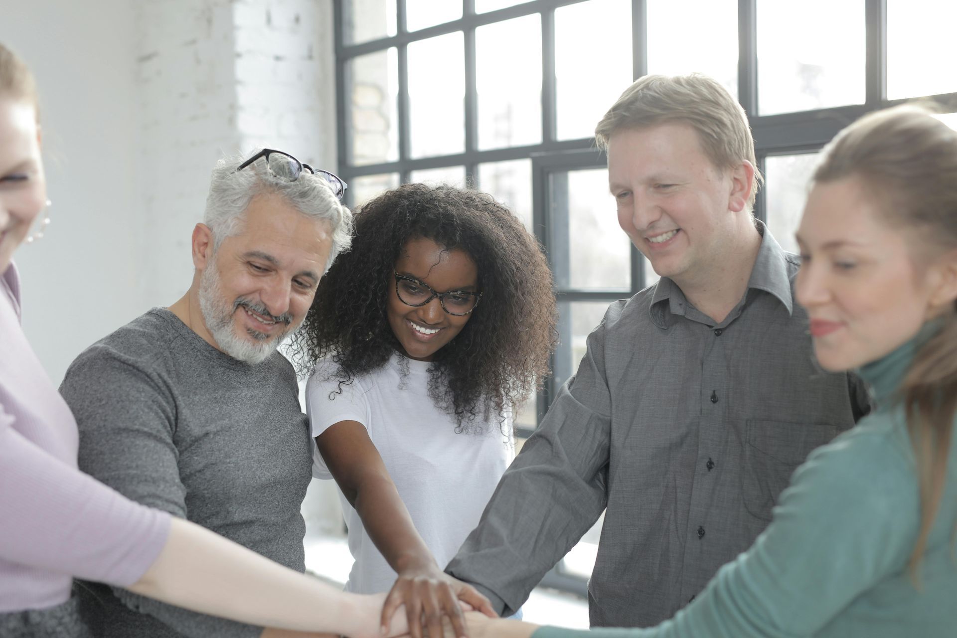 Group of people, smiling, with hands together in a circle, indoors by a window.