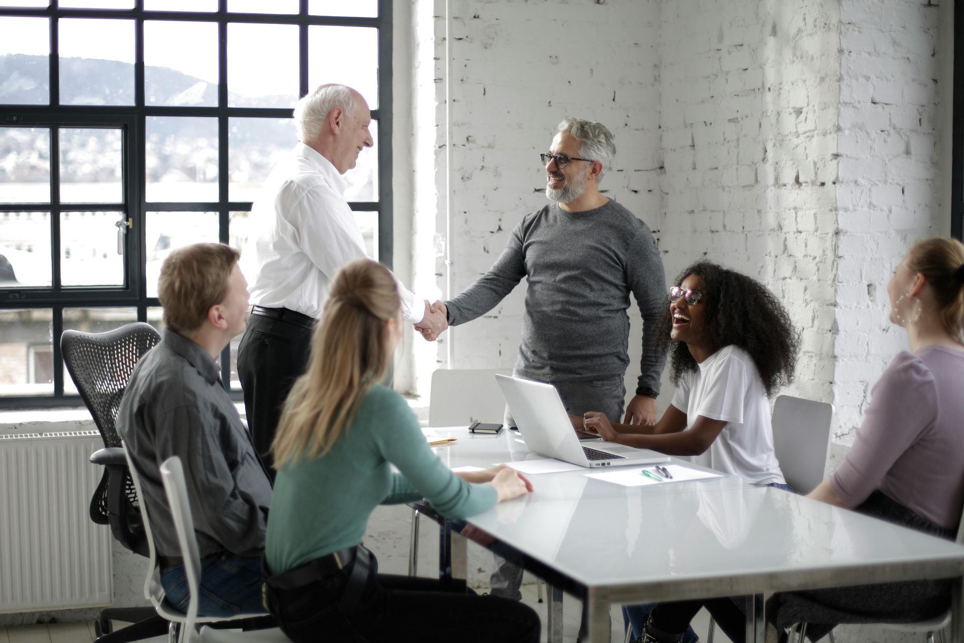 People in office, two men shaking hands. Others seated at a table. Laptop, window.