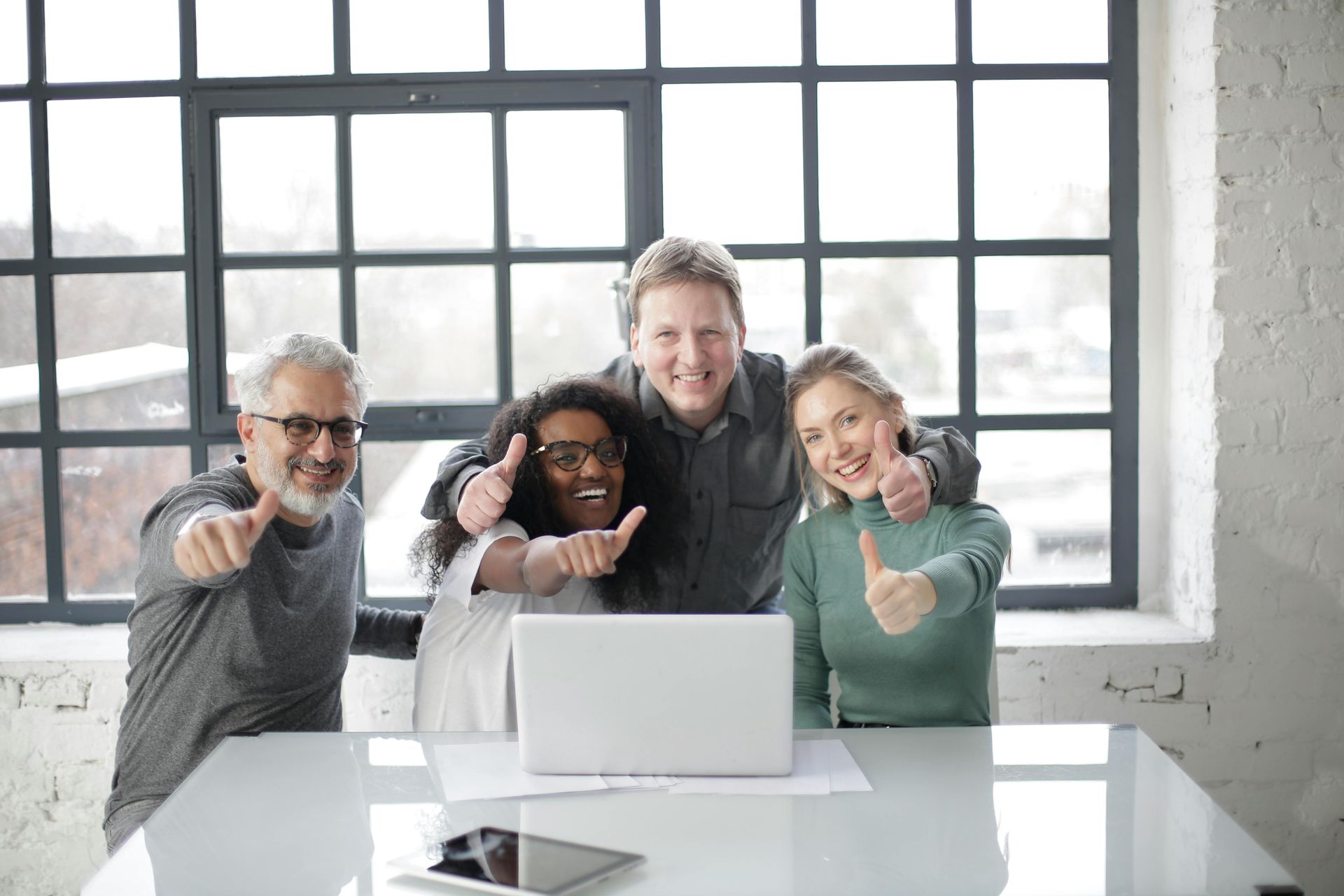 Four people in office, smiling, thumbs up by a laptop in front of window.