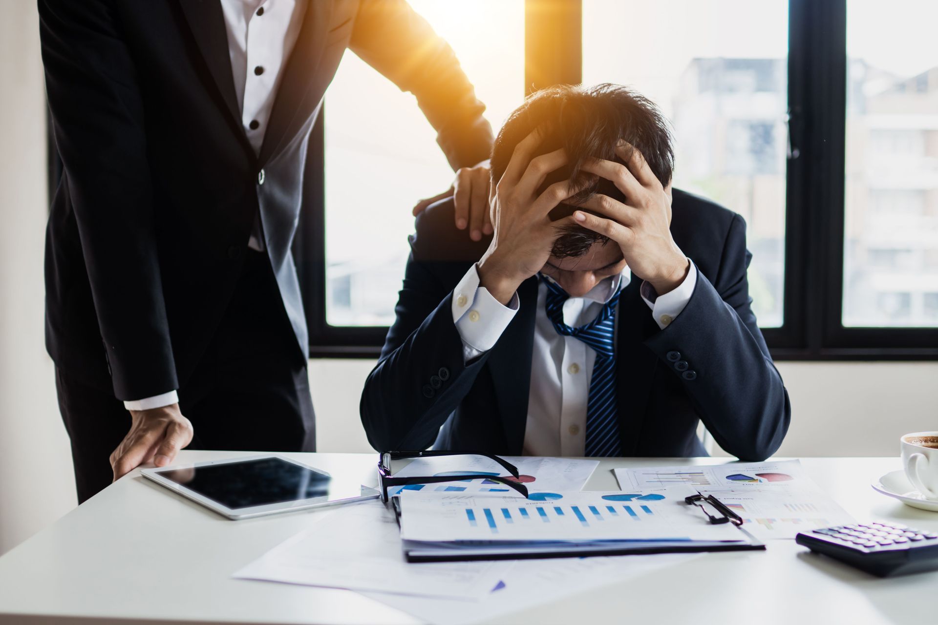 Businessperson stressed at desk while colleague comforts them in a bright office, with charts and tablet nearby