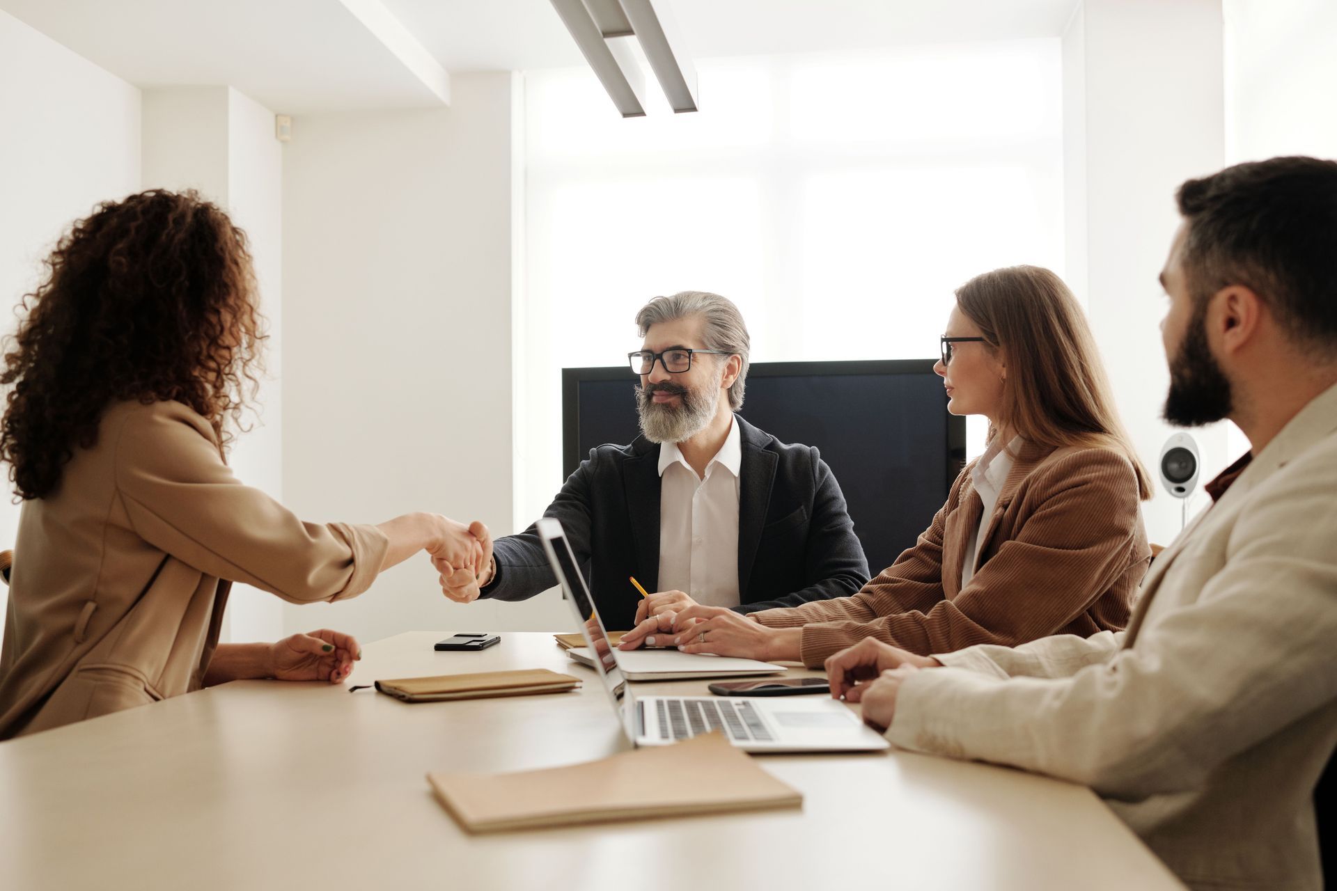 Four coworkers in a bright office shake hands during a meeting around a table.