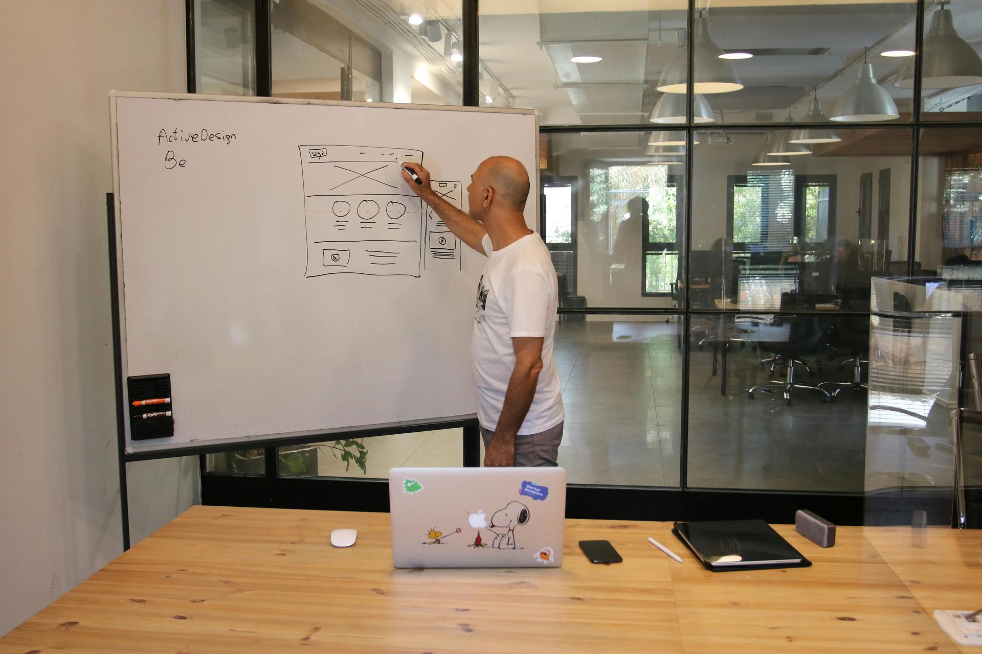 A person in a white t-shirt draws a diagram on a large whiteboard in a modern office with glass walls.
