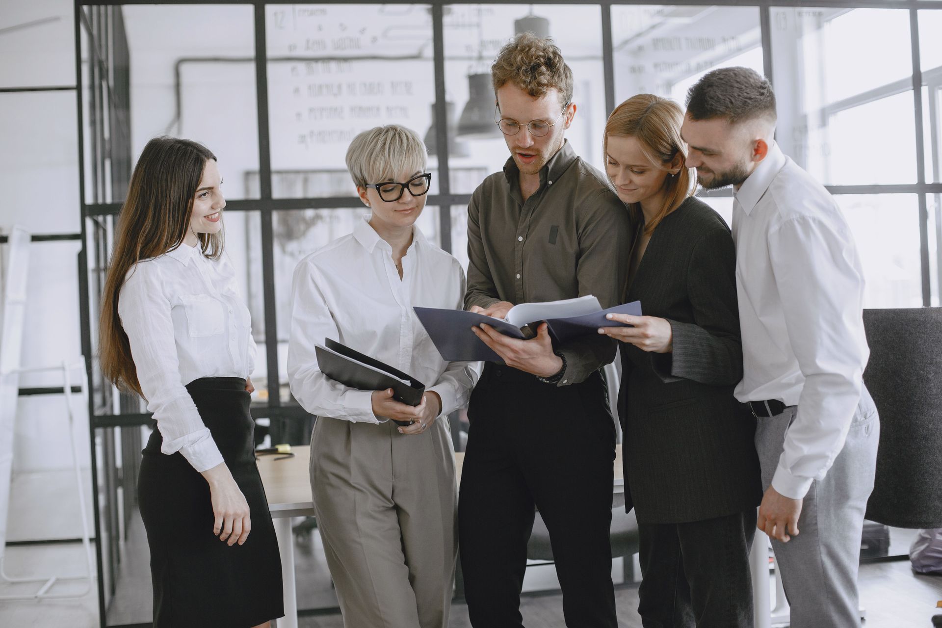 Five colleagues in business attire gather around a laptop in a bright office, discussing documents together