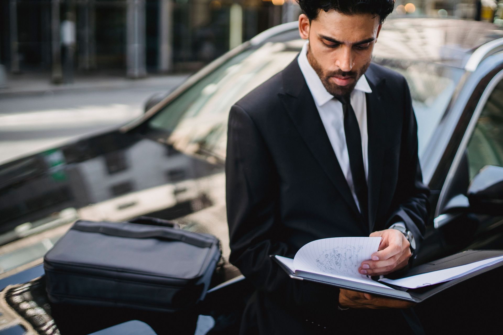 Man in a black suit reviewing documents beside a car on a city street