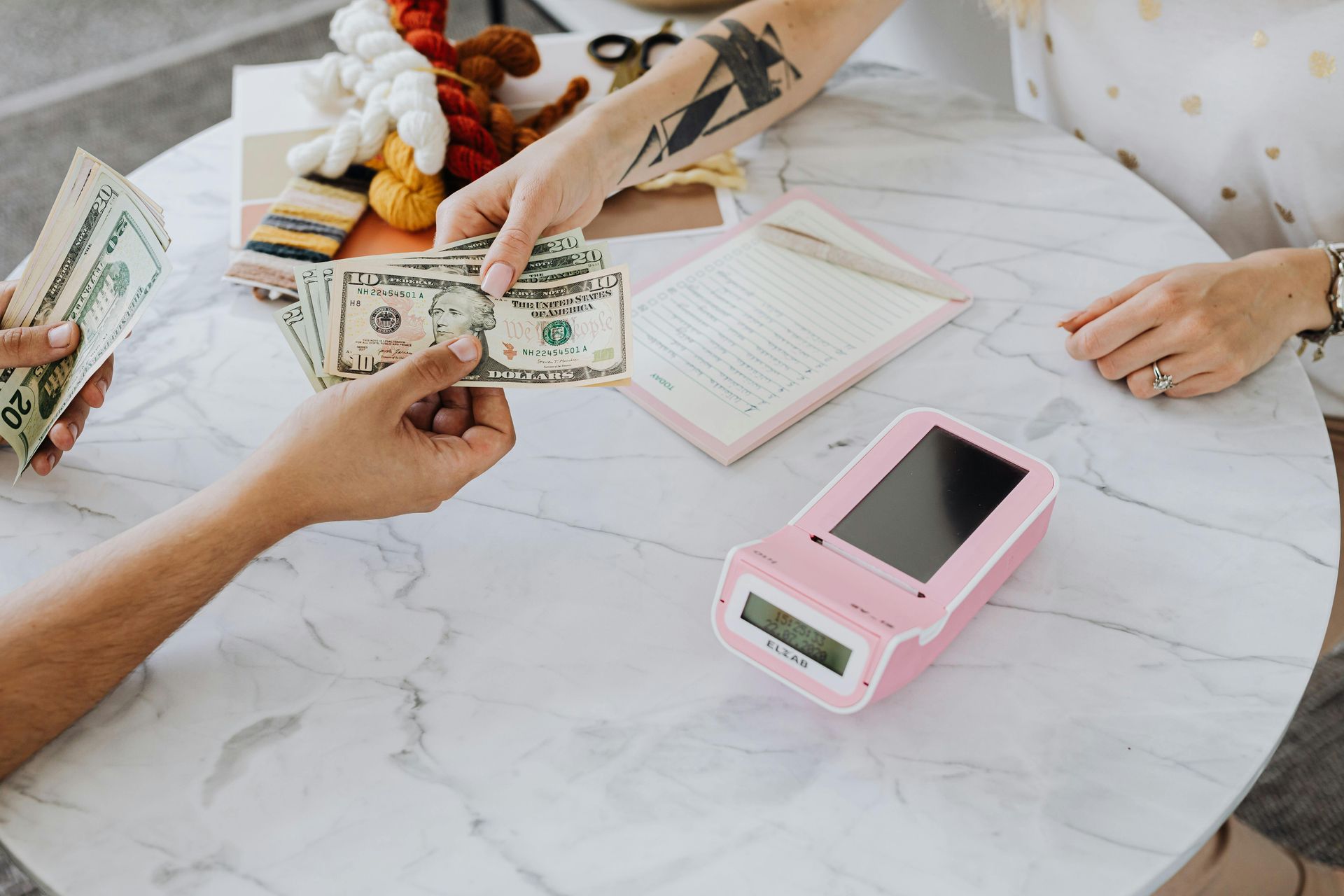 Two people exchange cash over a marble-topped table featuring a pink digital scale and craft supplies.