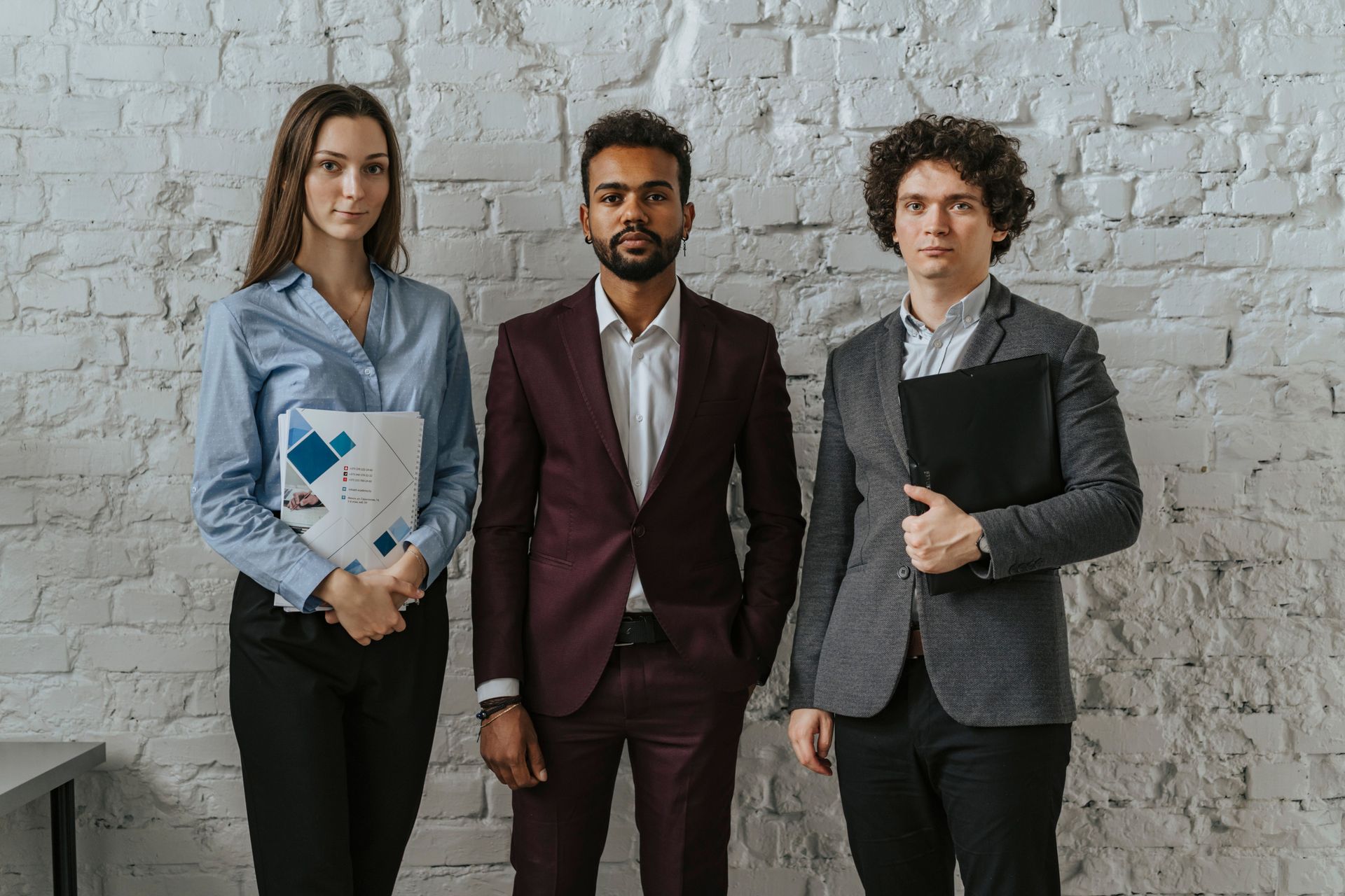 Three professionals stand against a white brick wall, holding folders and wearing business attire.