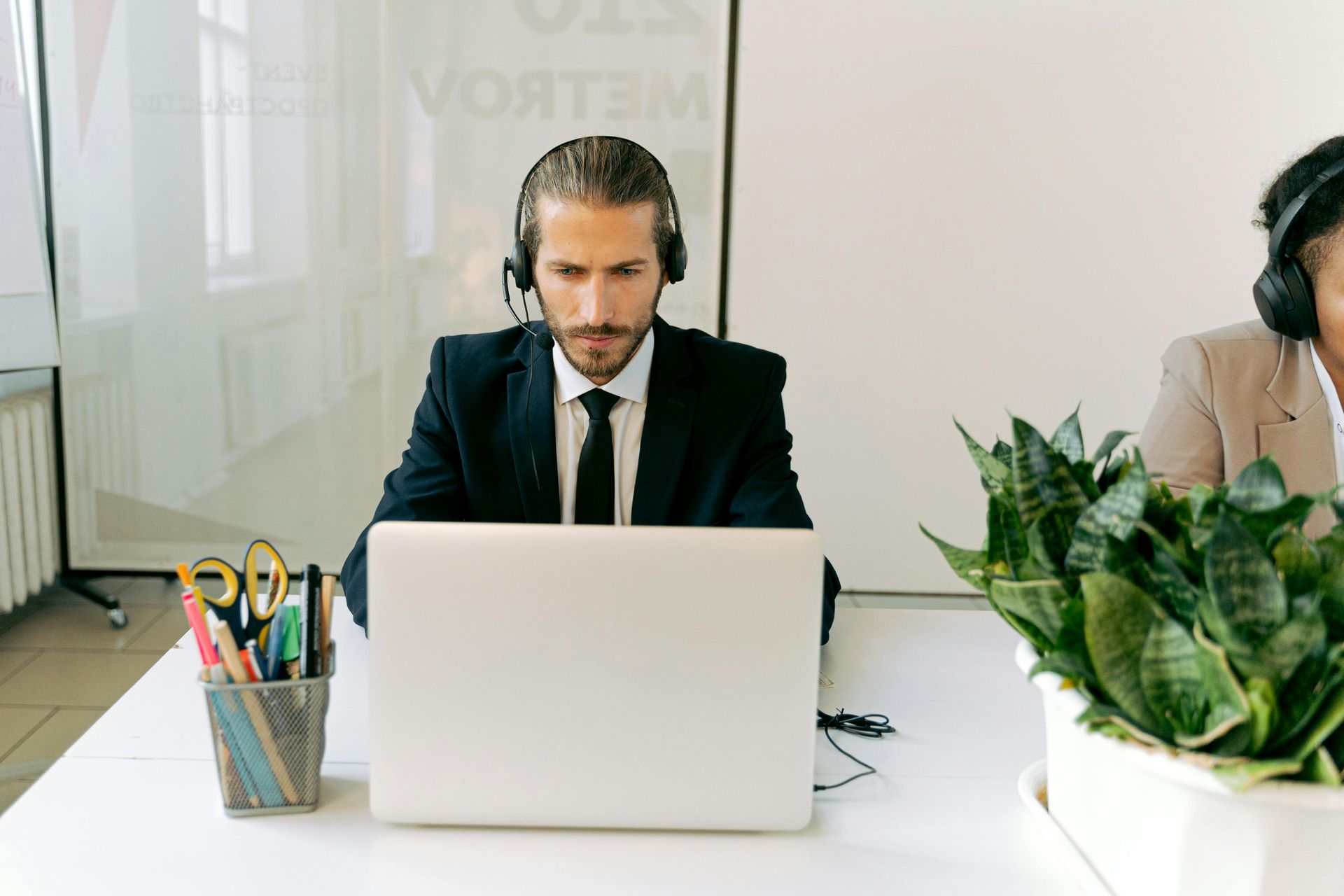 Man in a suit wearing headphones works on a laptop at a bright office desk with plants and pens.