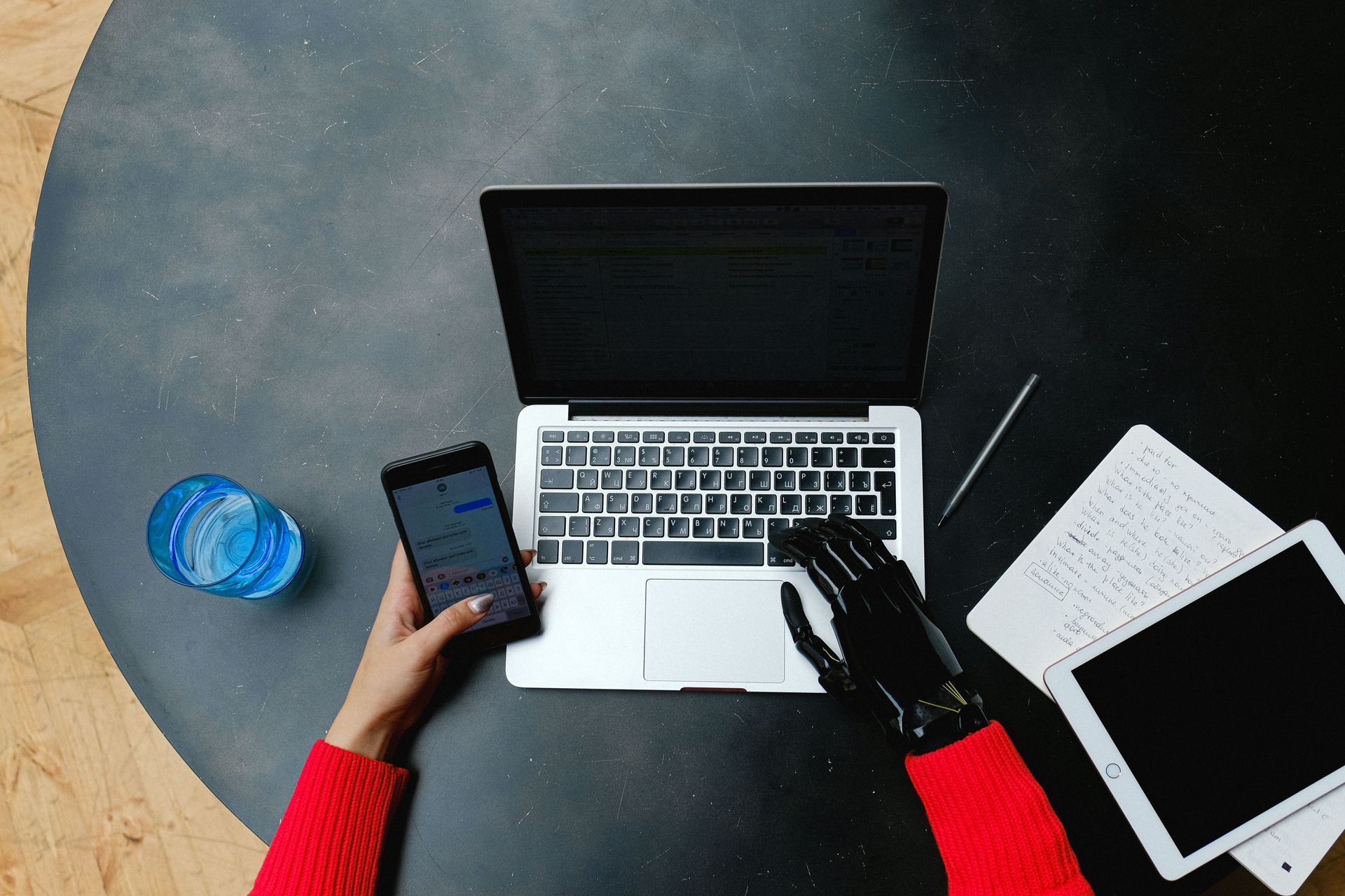 Hands typing on laptop beside smartphone, notebook, pen, and tablet on a round table