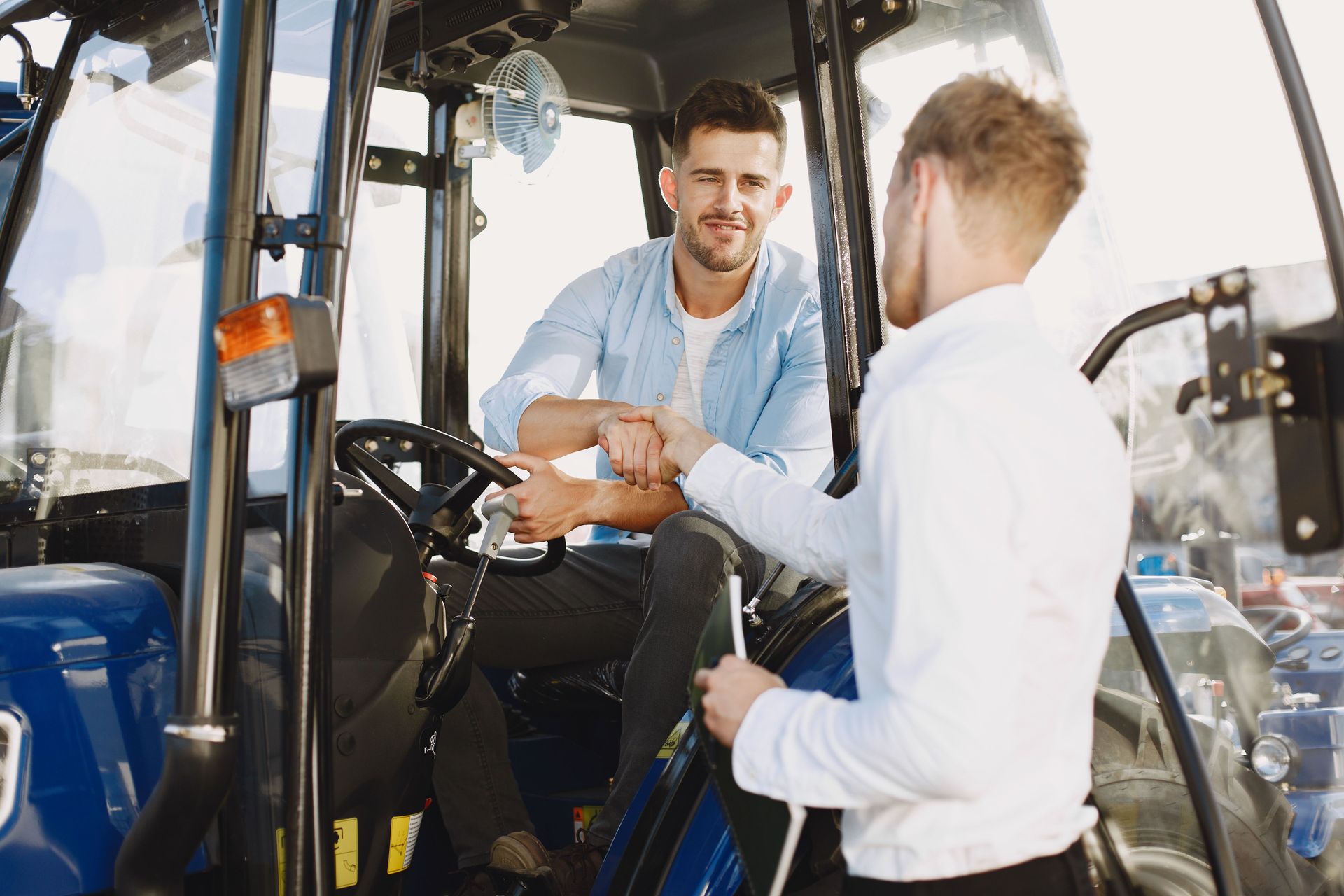 Two people shaking hands beside a blue tractor in a barn or workshop