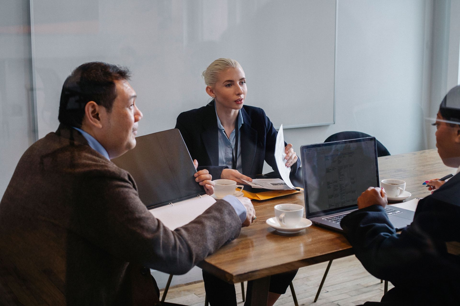 People meeting around a wooden table in a bright office, discussing documents and using laptops.