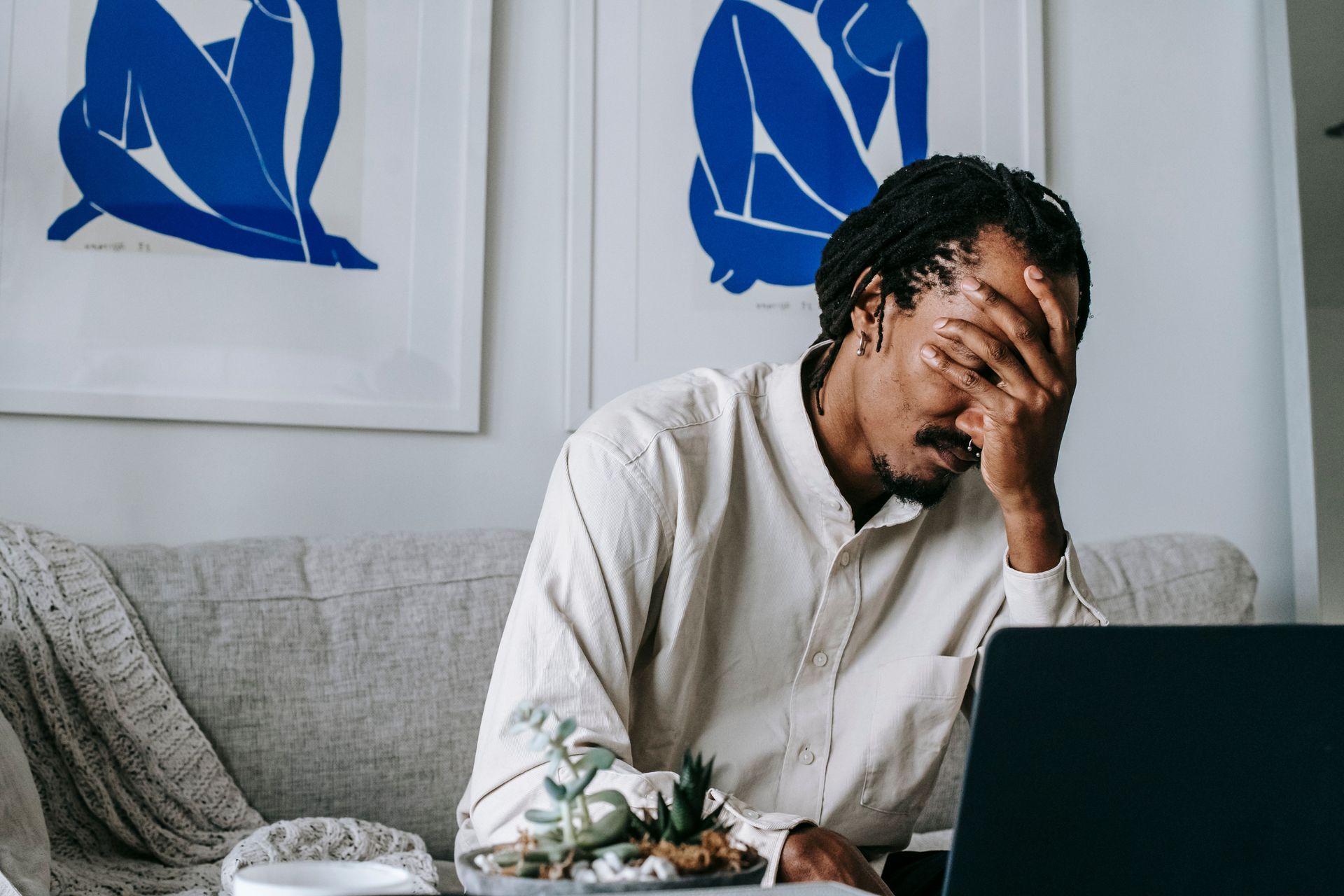 Person sits on a couch, hand on face, looking stressed beside an open laptop.