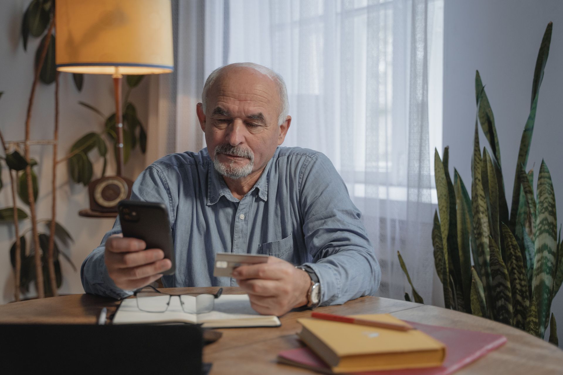 Man seated at a desk holding a phone and pen, with a lamp, notebooks, and plants in a home office.