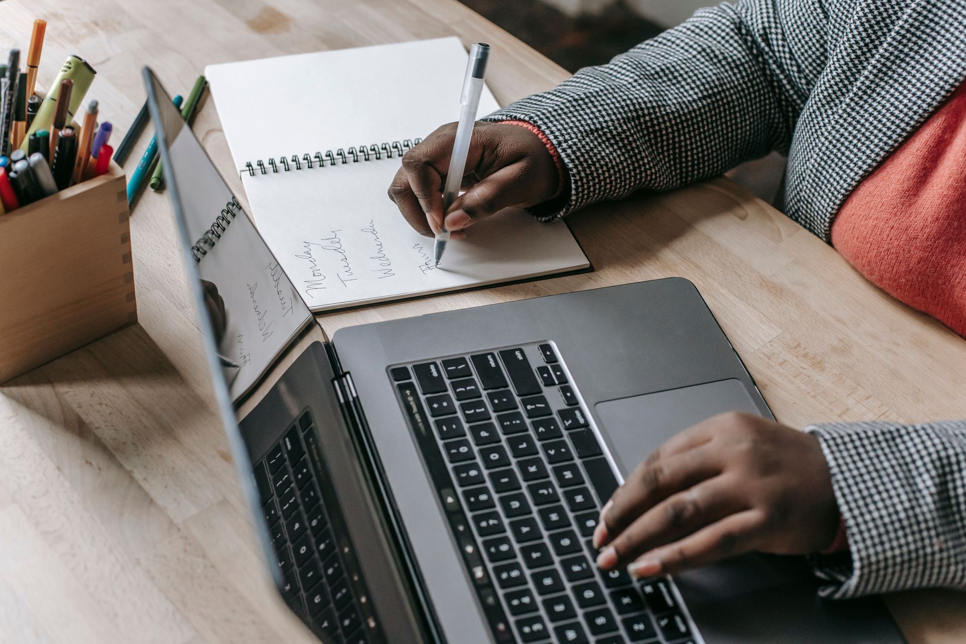 Person writing in a notebook beside a laptop at a desk with colorful pencils and papers