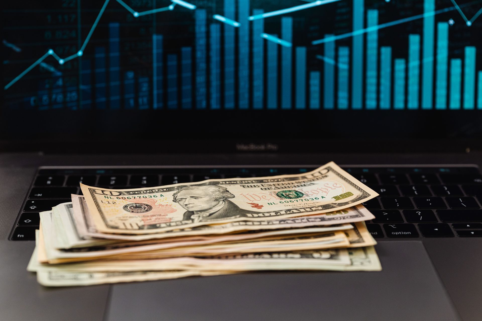 Stack of U.S. dollar bills on a laptop keyboard with a blue financial chart in the background