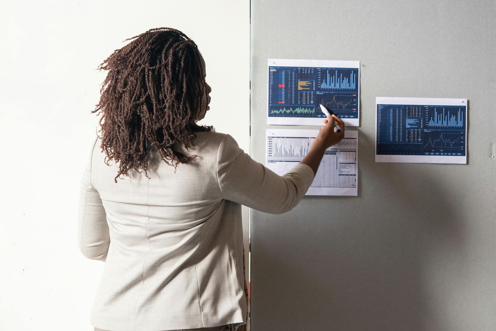 A person in a light blazer points to data charts attached to a light gray wall.