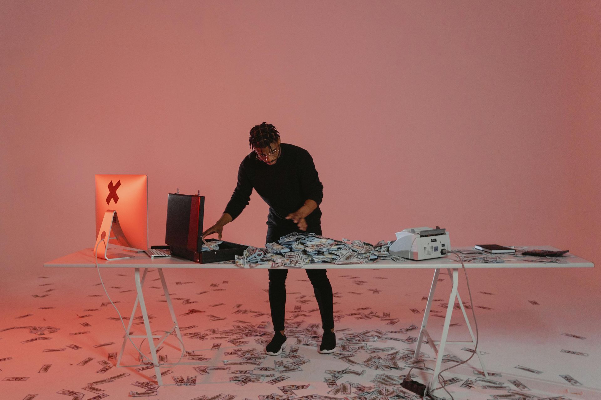 A person standing at a desk in a pink room, surrounded by scattered cash and office equipment, including a monitor.